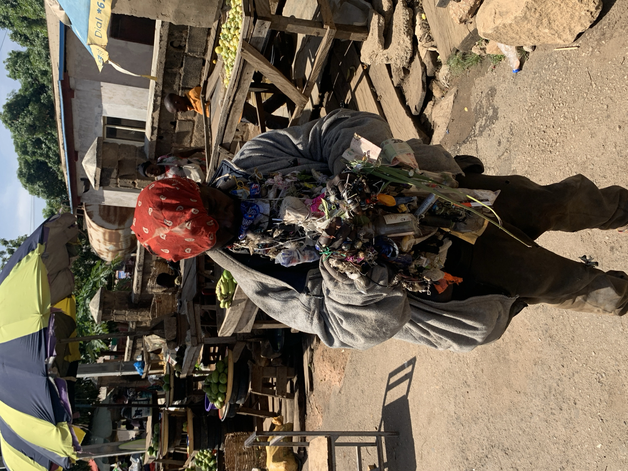 This image captures a vibrant street market scene in Jos, Nigeria, focusing on a street vendor. The central figure, seen from the waist up, wears a grey hooded sweatshirt and dark trousers, with a red patterned headscarf. This individual is prominently displaying a large, dense collection of various small goods, intricately attached to their upper body, likely for sale. These items include small plastic bags, bundles of green leafy herbs or spices, small bottles, trinkets, and other assorted merchandise. A banknote with the number "20" is visible among the items, suggesting local currency.
The vendor is either walking or standing on an uneven, light-colored dirt and gravel road. Strong sunlight casts clear shadows, indicating it is daytime, likely mid-morning or afternoon.
In the background, typical market infrastructure is visible: wooden stalls laden with green produce, possibly fruits like mangoes or plums. Other people, presumably market-goers or vendors, can be seen amidst the stalls. To the left, a large, two-toned yellow and blue striped umbrella provides shade. Simple concrete block buildings line the street, one featuring a blue tarp with "Dial *62" printed on it. Other visible text includes "Aquafina" and "FILMIGATO" on small packages among the vendor's wares. The overall setting is bustling and informal, characteristic of an open-air market.