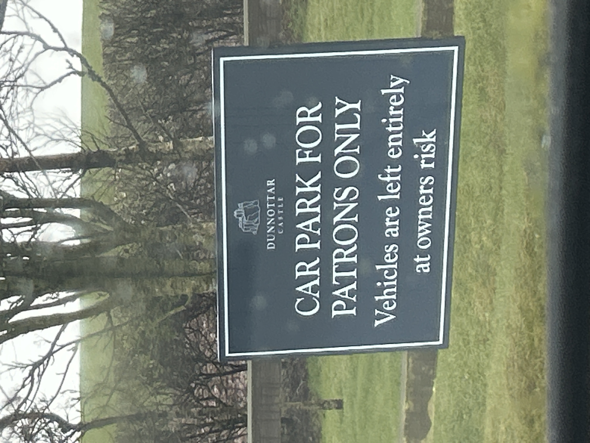 The image depicts a sign indicating the car park is for patrons only at Dunnottar Castle, located in Stonehaven, United Kingdom. The sign is dark with white lettering and a white border. Underneath "CAR PARK FOR" is the Dunnottar Castle logo. The sign also states "Vehicles are left entirely at owners risk." The sign is positioned outdoors, near what appears to be a tree without leaves and a grassy area. The image is somewhat obscured, possibly taken from inside a vehicle.