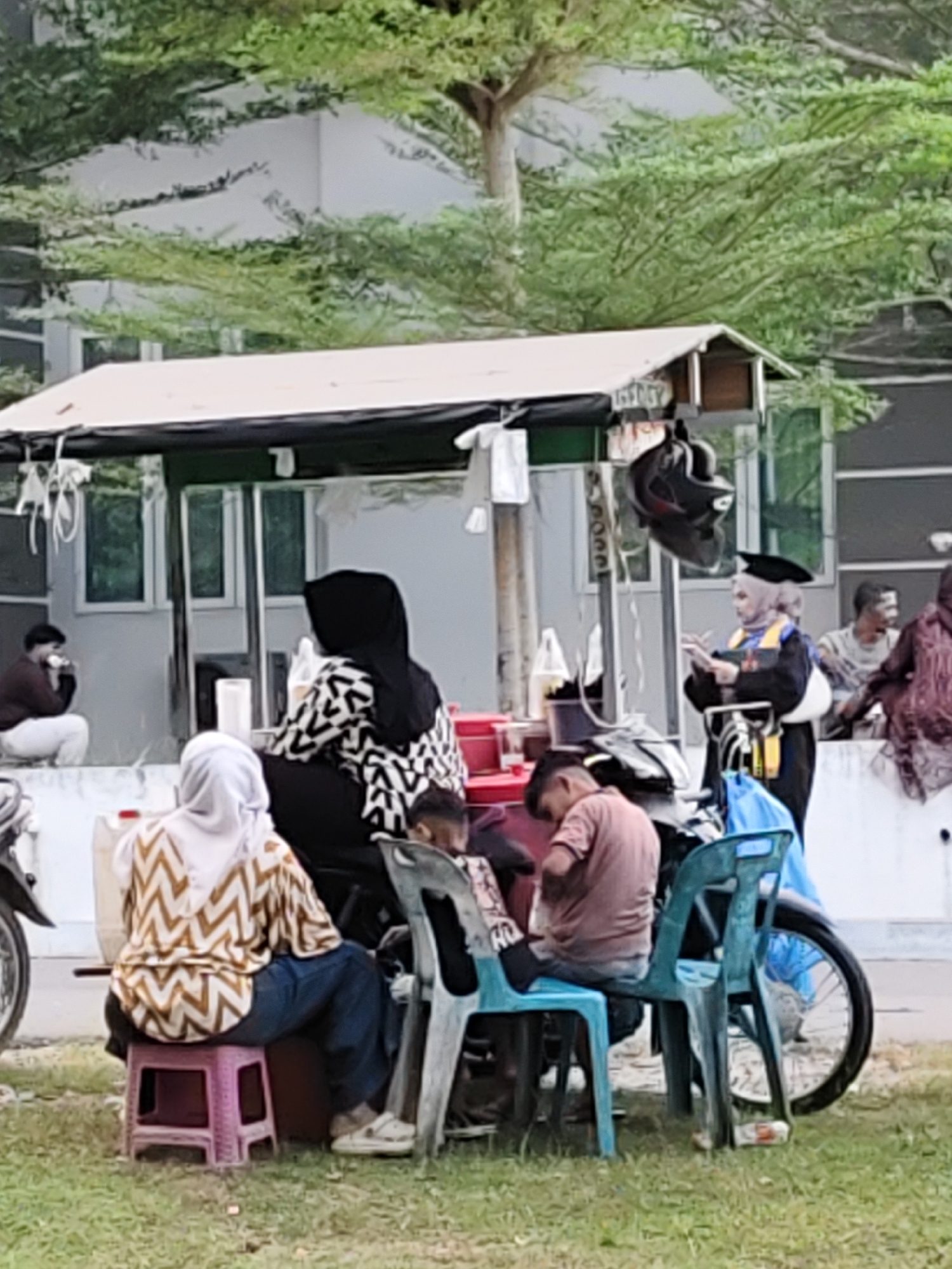 This image captures a lively outdoor scene in Lhokseumawe, Indonesia, featuring several individuals gathered around an informal vendor stall. In the foreground, a grassy patch leads up to a paved area where most of the activity takes place.
The central focus is a simple vendor stall with a white canopy and dark supports, suggesting a casual eatery or drink stand. Red plastic containers are visible on the stall's counter, and a black motorcycle helmet hangs from one of its beams.
Several people are seated in front of and around the stall. To the left, a woman wearing a white hijab and a brown and cream patterned top sits on a pink plastic stool, with her back to the viewer. Adjacent to her, another woman in a black hijab and a black and white patterned top appears to be tending to the stall or a child seated nearby. Two young boys are also present, one in a brown t-shirt sitting on a light blue plastic chair, and another child's head visible closer to the black-hijab-wearing woman. A young man in a light brown t-shirt sits on a dark green plastic chair on the right side of this group, leaning forward. A motorcycle wheel is visible on the far left and right edges of the frame.
Further to the right, a woman stands out, dressed in a black graduation gown with yellow or gold trim and a mortarboard cap over a light-colored hijab. She is holding what appears to be a book or tablet and looking towards the right, possibly interacting with someone out of frame or observing the surroundings.
In the background, behind a low white wall, a white building with multiple windows is visible. Trees with lush green foliage frame the top and background of the image, indicating an open, somewhat green setting. Other individuals are seen sitting on the white wall in the background; one person on the left appears to be eating or drinking, and two men are seated on the right side of the wall.
The scene depicts a relaxed daytime gathering, possibly a post-graduation celebration or a casual social interaction at a local vendor spot. The overall atmosphere is informal and communal.