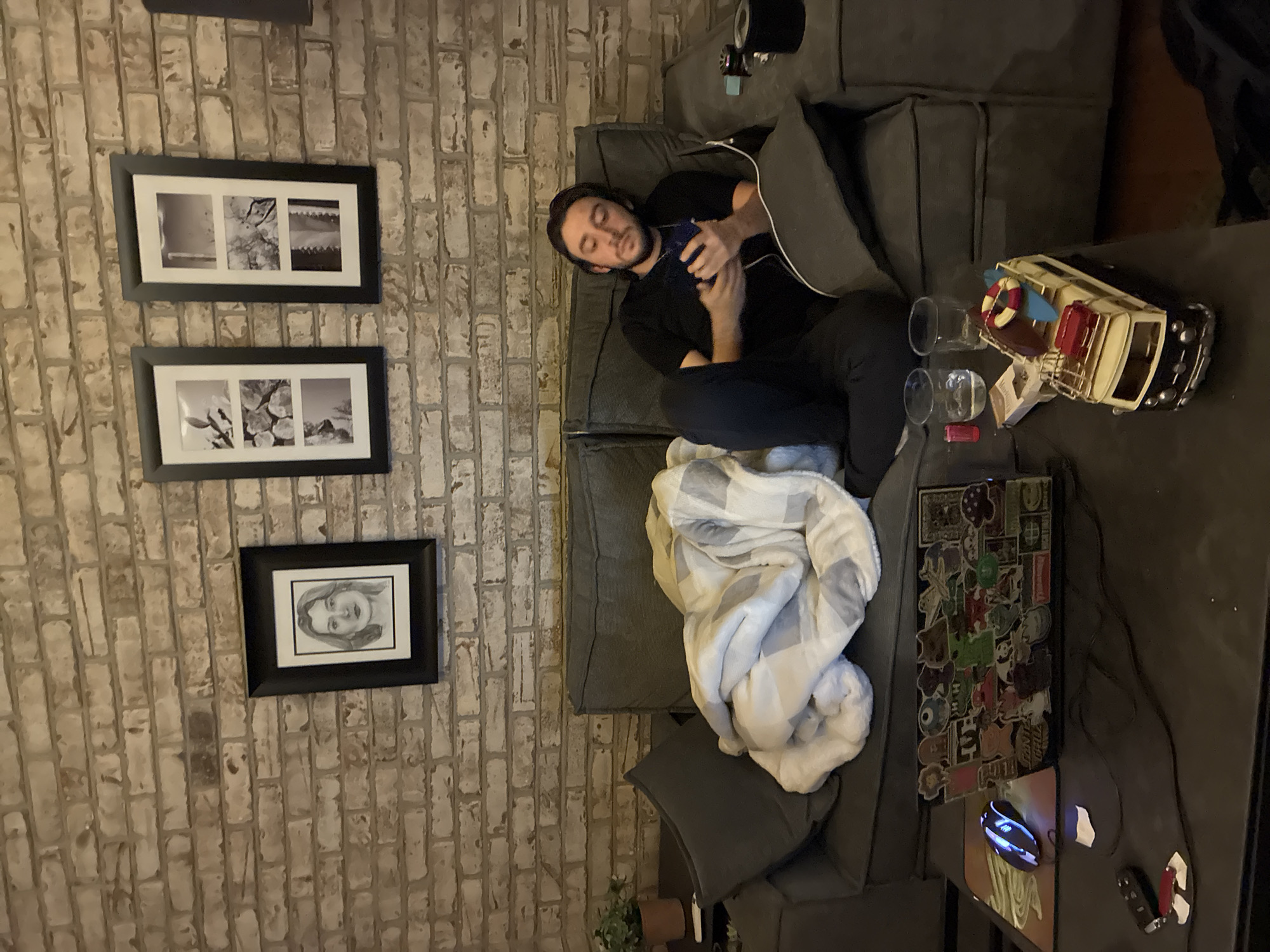 A person lies on a gray couch with a blanket. They are using a phone. The couch is next to a brick wall adorned with three framed pictures. On the table next to the couch, there is a model of a boat, two glasses, a laptop with stickers, a mouse, and some other small objects. The location is an indoor setting, not Huixquilucan de Degollado, Mexico.