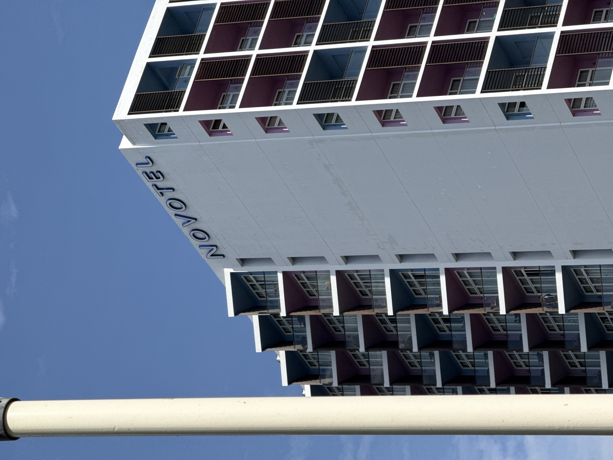 The image captures a close-up, low-angle view of the Novotel Nha Trang hotel, a modern high-rise building situated in Nha Trang, Vietnam. The hotel's facade is a prominent feature, with its white exterior adorned with rows of balconies. These balconies alternate in color between vibrant purple and cool blue, creating a striking visual pattern. Many of the balconies feature glass railings and sliding doors leading to windows, suggesting individual hotel rooms. The hotel's name, "NOVOTEL," is prominently displayed in blue lettering across the upper section of the building.
To the left of the hotel, a tall, cylindrical, cream-colored structure, likely a pillar or a part of a larger edifice, occupies a significant portion of the frame. This structure is slightly out of focus, drawing the viewer's attention to the hotel's architecture.
The background consists of a clear, bright blue sky with a few wispy white clouds. The overall lighting suggests a sunny day. There are no visible people or activity in the image, contributing to a still and architectural focus. The scene is framed from a perspective that emphasizes the building's height and repetitive design elements.