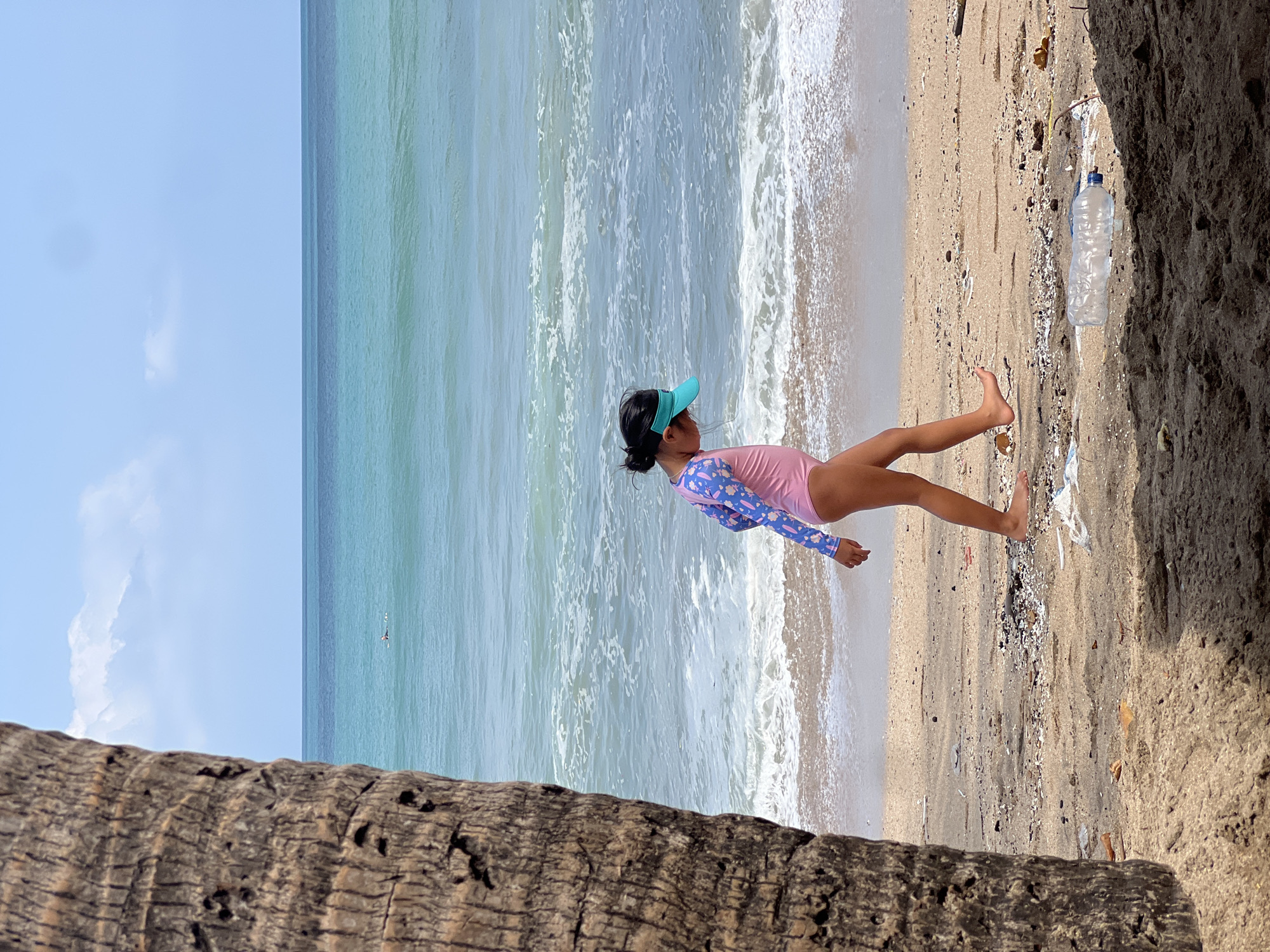 A young girl wearing a pink swimsuit with blue sleeves and a teal visor walks along a sandy beach in Kuta, Indonesia. The ocean, with turquoise water and white waves, is to her right. In the foreground, a portion of a palm tree trunk is visible on the left, and debris, including a plastic bottle, is scattered on the sand. The sky is a clear blue with a few white clouds.