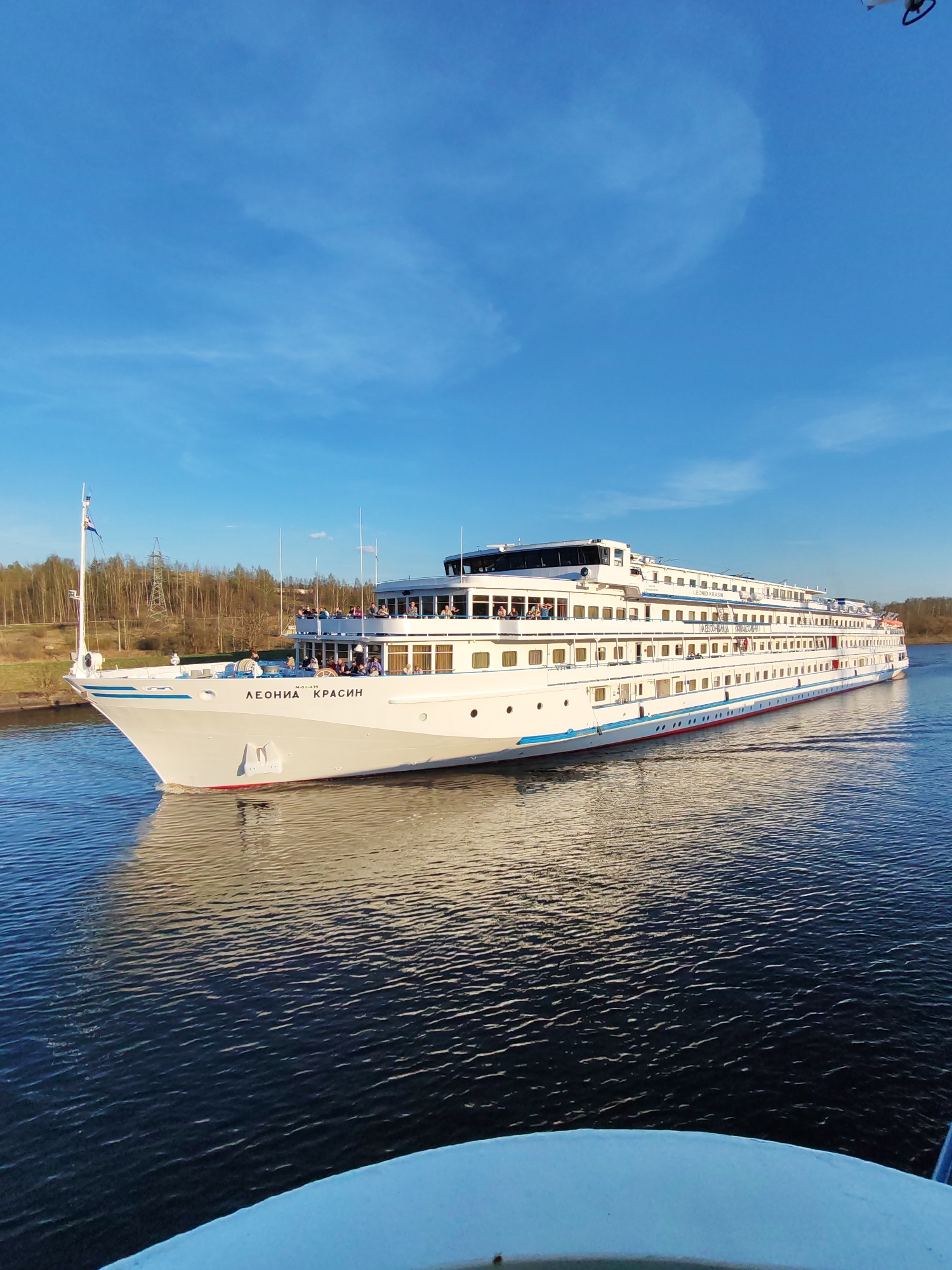 A long, white cruise ship, named "Леонид Красин", is sailing on a body of water. The sky is a clear blue with a few wispy clouds. Trees and vegetation are visible on the shore in the background. The location is near Базарово, Russia, as evidenced by the presence of power lines that are visible through the trees. People are seen on the cruise ship. The foreground shows a portion of another vessel's white railing or deck.