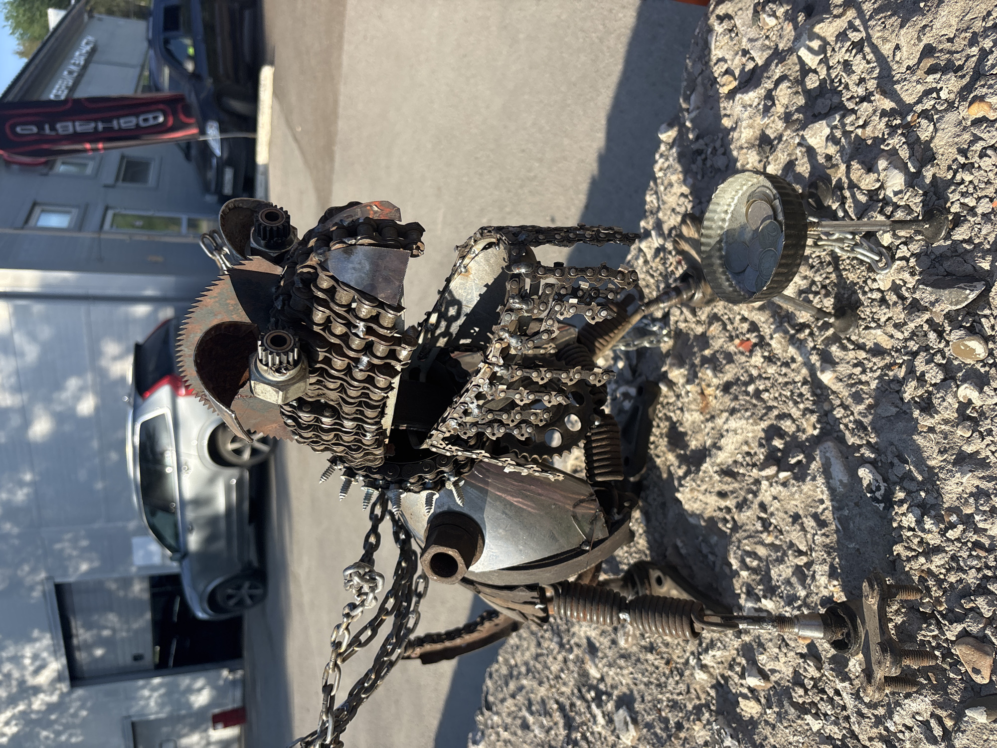 The image shows a sculpture made of scrap metal, resembling an animal figure, placed on a rough, rocky surface. The sculpture appears to be constructed from various mechanical parts such as chains, gears, saw blades, nuts, bolts, springs, and metal sheets. The sculpture's head is formed by the circular saw blade, and chains form a part of its body. The background shows a street with a grey car parked near a building with a sign “ФАВТО Автосервис”. The location is likely Perm, Russia, given the Cyrillic writing on the sign. The scene is outdoors in daylight.