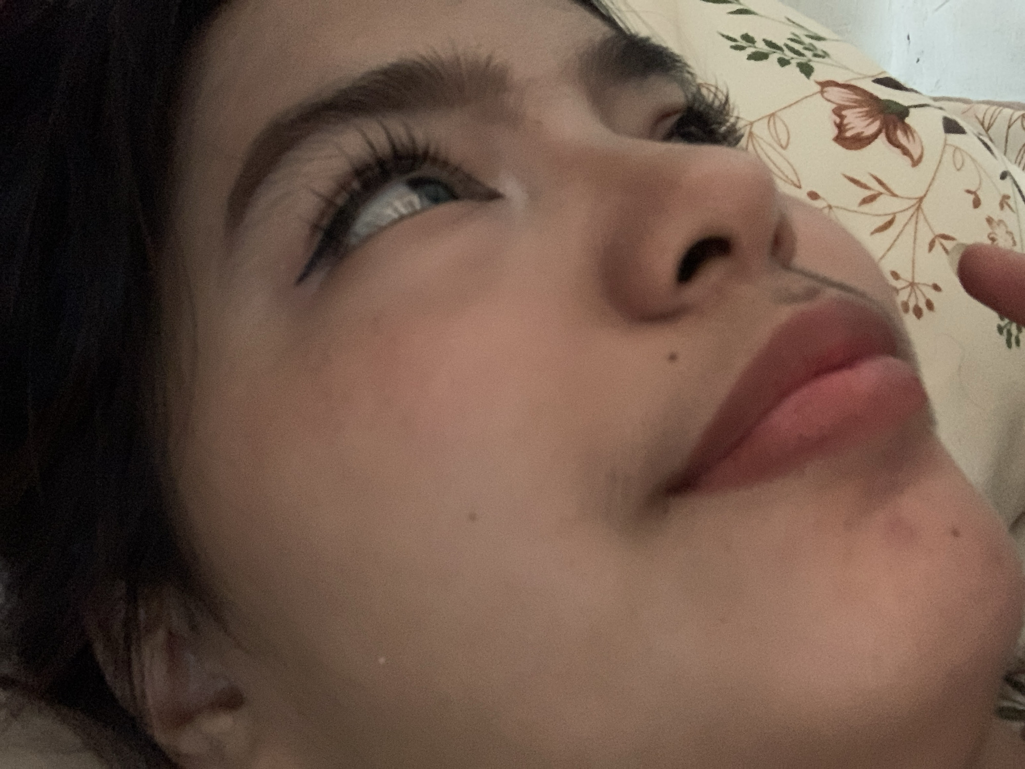 A close-up, angled shot captures the right side of a person's face, likely female, from a low perspective. The person is looking upwards and to the left of the frame. Her right eye is prominent, featuring a light-colored iris, distinct eyelashes, and a subtle winged eyeliner. Her eyebrow is dark and well-defined. The bridge of her nose, her upper lip, and a portion of her lower lip and chin are also visible. A small mole or beauty mark is present on her cheek and another on her chin. Her skin appears smooth.
In the background, on the right side of the image, a light-colored pillow with a brown and green floral pattern (showing motifs of flowers and leaves) is visible, suggesting an indoor setting, possibly a bed or sofa. A finger, presumably belonging to the subject, lightly touches the area near the corner of her mouth on the far right.
The lighting is soft, creating gentle shadows and highlights on her face. The overall mood appears calm and relaxed, with the subject seeming to be in a thoughtful or introspective state. No text is visible in the image.