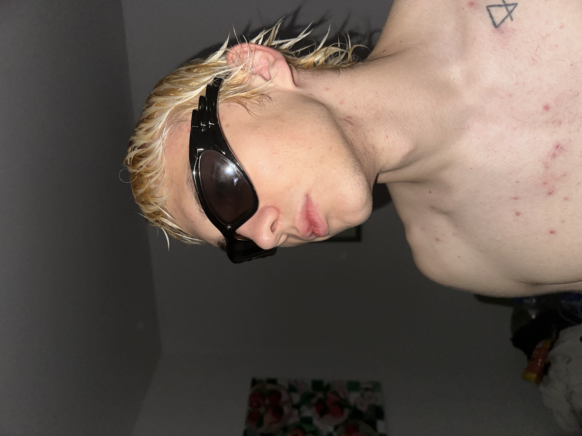 The image shows a person with short, blonde, wet hair and wearing black sunglasses. The person's bare chest is visible, showing some skin blemishes. A tattoo of a triangular symbol is located on the upper chest. The background is dark, with a blurry floral design on the wall. The image appears to be taken indoors. There is no information in the image to determine if this image was taken in the city of Novi Sad, Serbia.