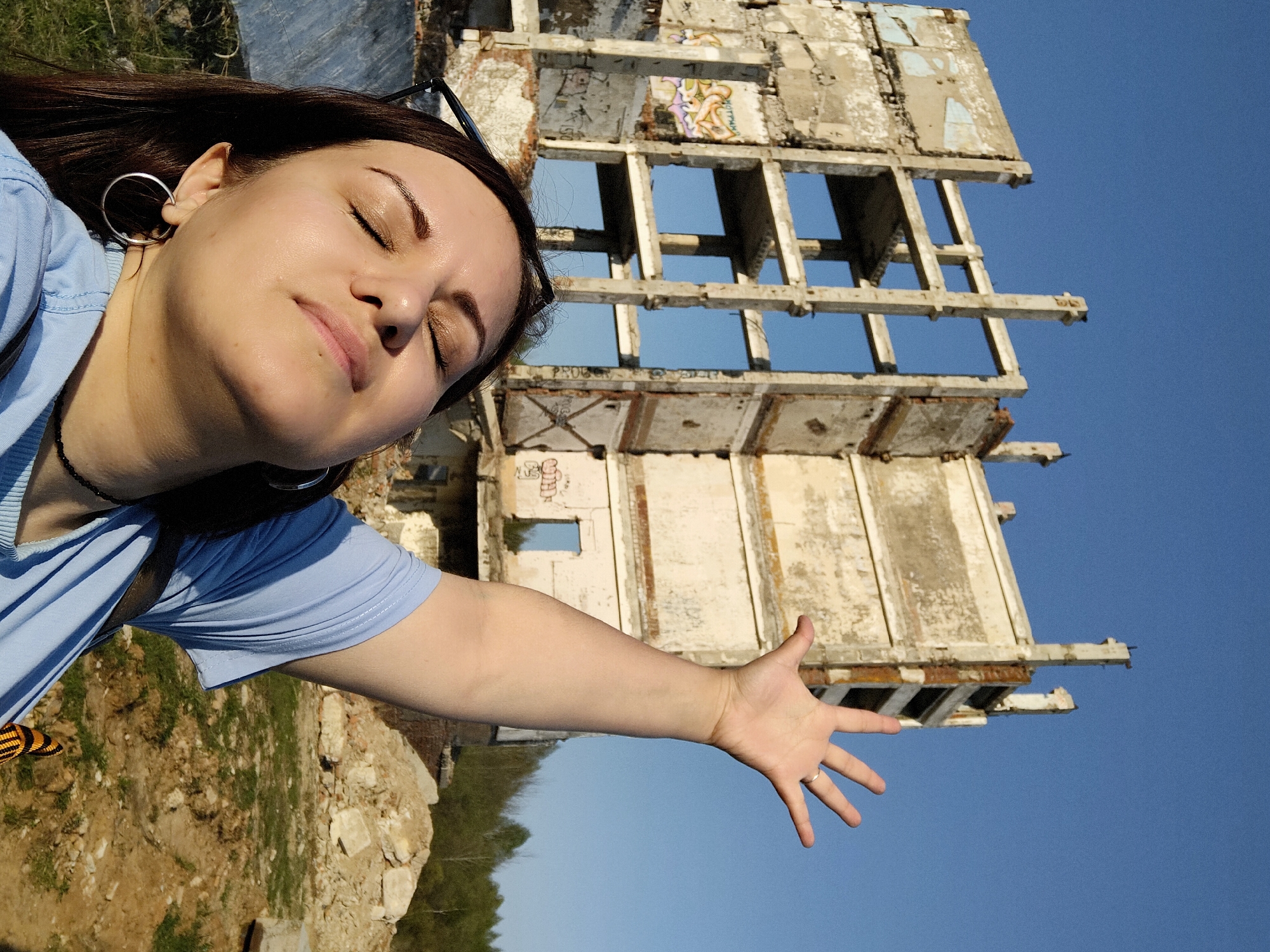A low-angle medium shot depicts a woman with dark hair and fair skin, positioned on the left, extending her right arm towards the right side of the frame. Her eyes are closed, and she appears to be facing the sun. She wears silver hoop earrings, a blue top, and a black beaded necklace.
In the background stands a partially ruined, multi-story building made of concrete and white paint, with numerous empty window frames. The building is in disrepair with graffiti visible on some of the walls. The sky behind the building is clear and blue.
Below the building, in the lower left corner, there is a glimpse of a grassy area with some rocks and debris, possibly indicating the ground level.
Based on the information provided, the location is Марковский, Russia.
