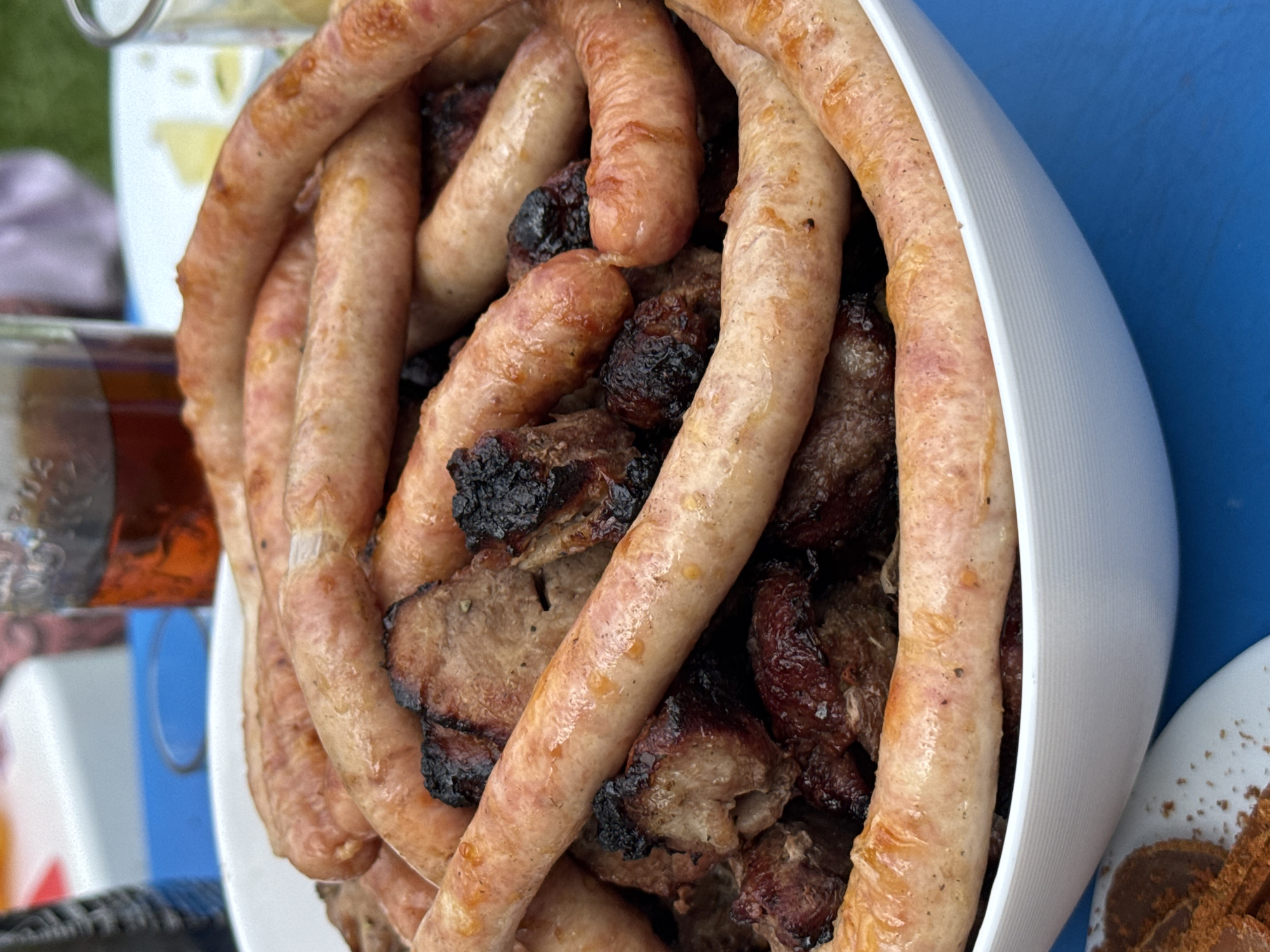 A close-up, overhead shot shows a white bowl filled with grilled sausages and chunks of meat. The sausages, which are light brown and plump, are arranged in a circular pattern on top of the meat. The meat, appearing to be pork, is dark brown and has some charred edges. The bowl is placed on a bright blue surface, likely a table, and a blurred background includes a glass of amber liquid and hints of green grass. The lighting suggests an outdoor setting, possibly a backyard barbecue.