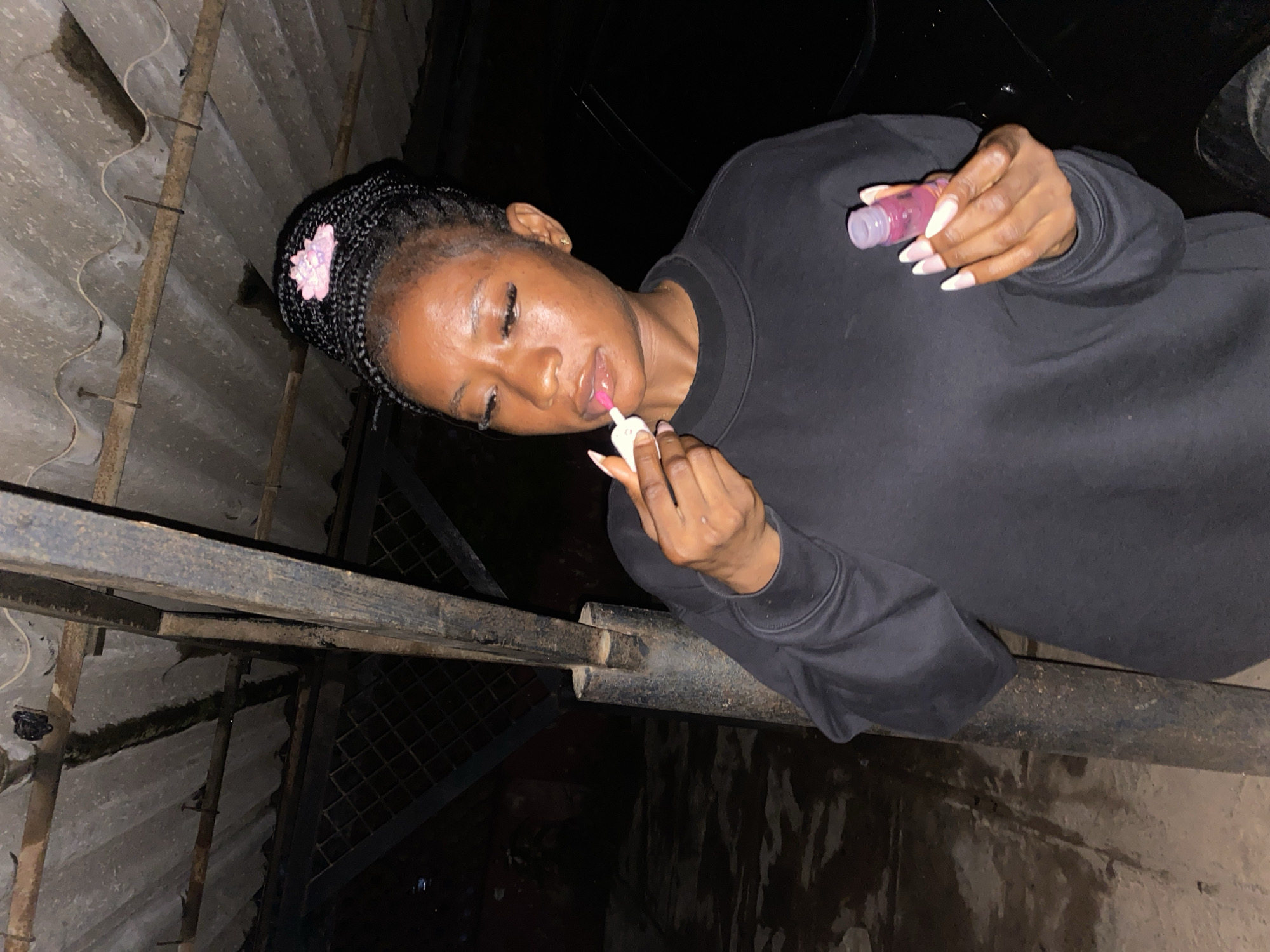 A young Black woman with braided hair adorned with a pink accessory is applying lip gloss in what appears to be an outdoor, covered area, possibly a carport or shelter, at night. She is wearing a black sweatshirt. In her right hand, she holds the lip gloss wand to her lips, and in her left hand, she holds the lip gloss container. Her nails are long and painted with a French manicure. The background is dark and indistinct, with hints of rough textures and a metal structure overhead. The scene is likely in Jos, Nigeria, given the prompt's context.