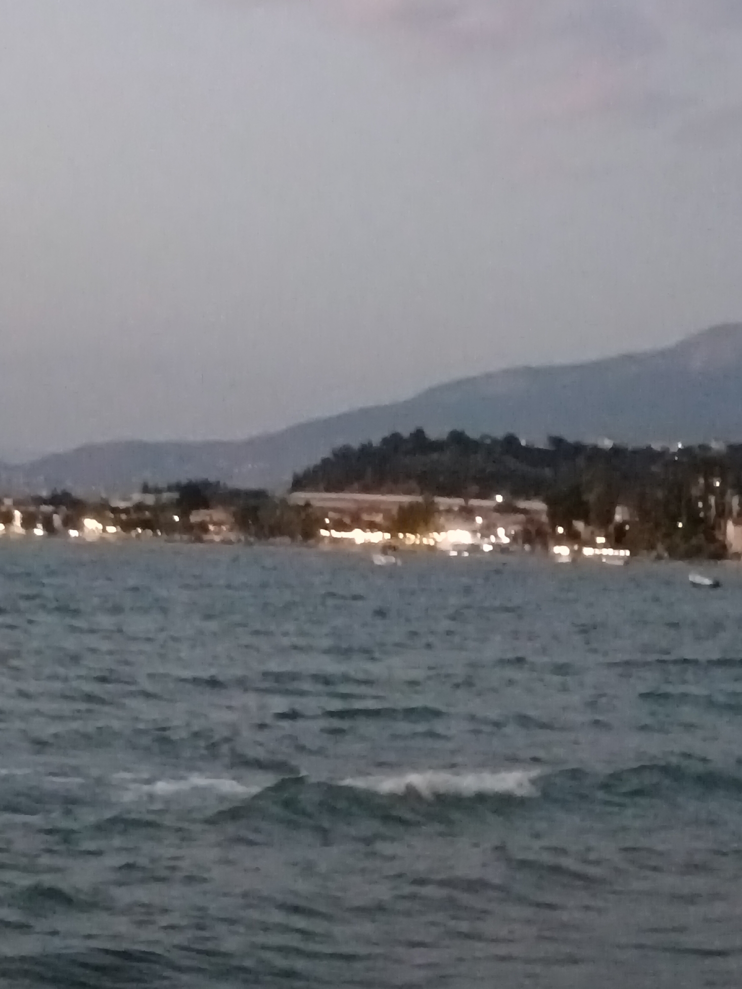 The image depicts a coastal scene at dusk or dawn. In the foreground, a body of water with gentle waves is visible. The water appears dark and rippled. In the midground, a shoreline is lined with buildings and illuminated by numerous lights, suggesting a populated area. Trees and vegetation are interspersed along the coastline, and hills or mountains form a backdrop to the settlement. A few small boats are discernible on the water, one to the right and others further left. The sky is overcast with a muted light. The location appears to be a coastal town or resort area within the Municipal Unit of Vrachneika, Greece, given the natural landscape and the presence of illuminated buildings along the water.