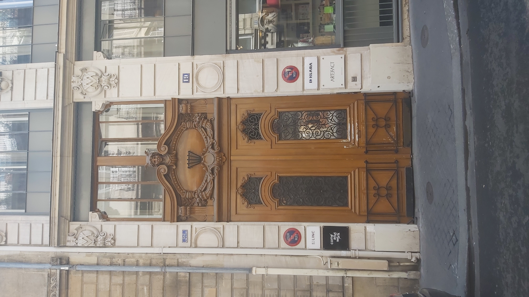 A medium shot captures the ornate entrance of a building on a Parisian street. The central focus is a large, double wooden door with intricate carvings. The door is flanked by decorative columns and topped with an elaborate pediment featuring a sculpted face, possibly of Mercury due to the winged helmet. Above the doors, a large window with multiple panes reflects the surrounding buildings and sky.
To the left of the entrance, a vertical pipe runs down the facade, which is constructed of pale stone blocks. Several signs are affixed to the wall: a small blue sign labeled "GAZ" (gas) with an arrow, a circular red and blue sign that reads "JOUR ET NUIT" (day and night), a white sign with the text ":: LINC", and a black sign with white text that says "come and work".
To the right of the entrance, a light-colored column is adorned with decorative plasterwork and a house number "19" in blue on a white plaque. Below the number, there are two white signs, one reading "IO KLARA" and the other "ARTEFACT". Further to the right, a large shop window reveals shelves stocked with various items, suggesting a commercial space.
The ground in front of the building is paved with cobblestones, and a street with a curb is visible in the foreground. The overall impression is one of an established, possibly historic, building in an urban setting.