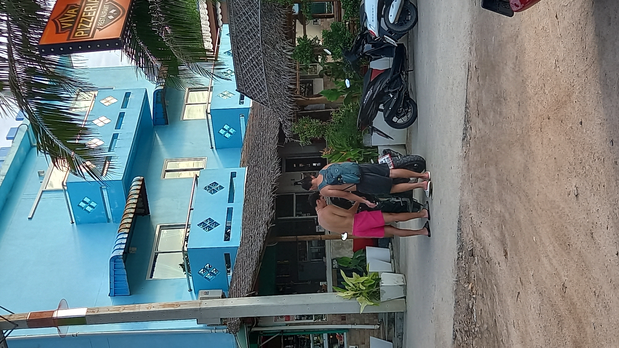 Two young men are standing next to a black motorcycle in front of a blue building with a thatched roof structure extending outwards. The man on the left is wearing pink shorts and is shirtless, while the man on the right is wearing dark shorts and a backpack, also shirtless. They appear to be interacting with the motorcycle. The building behind them has multiple balconies with decorative diamond patterns and a sign for "Viva Pizzeria Restaurant" is visible. Palm trees are present in the background, and a power pole is on the left side of the image. The ground is a dirt and paved road. This scene is consistent with the common streetscape found in ตำบลเกาะพะงัน, Thailand, a popular tourist destination known for its beaches and relaxed atmosphere.