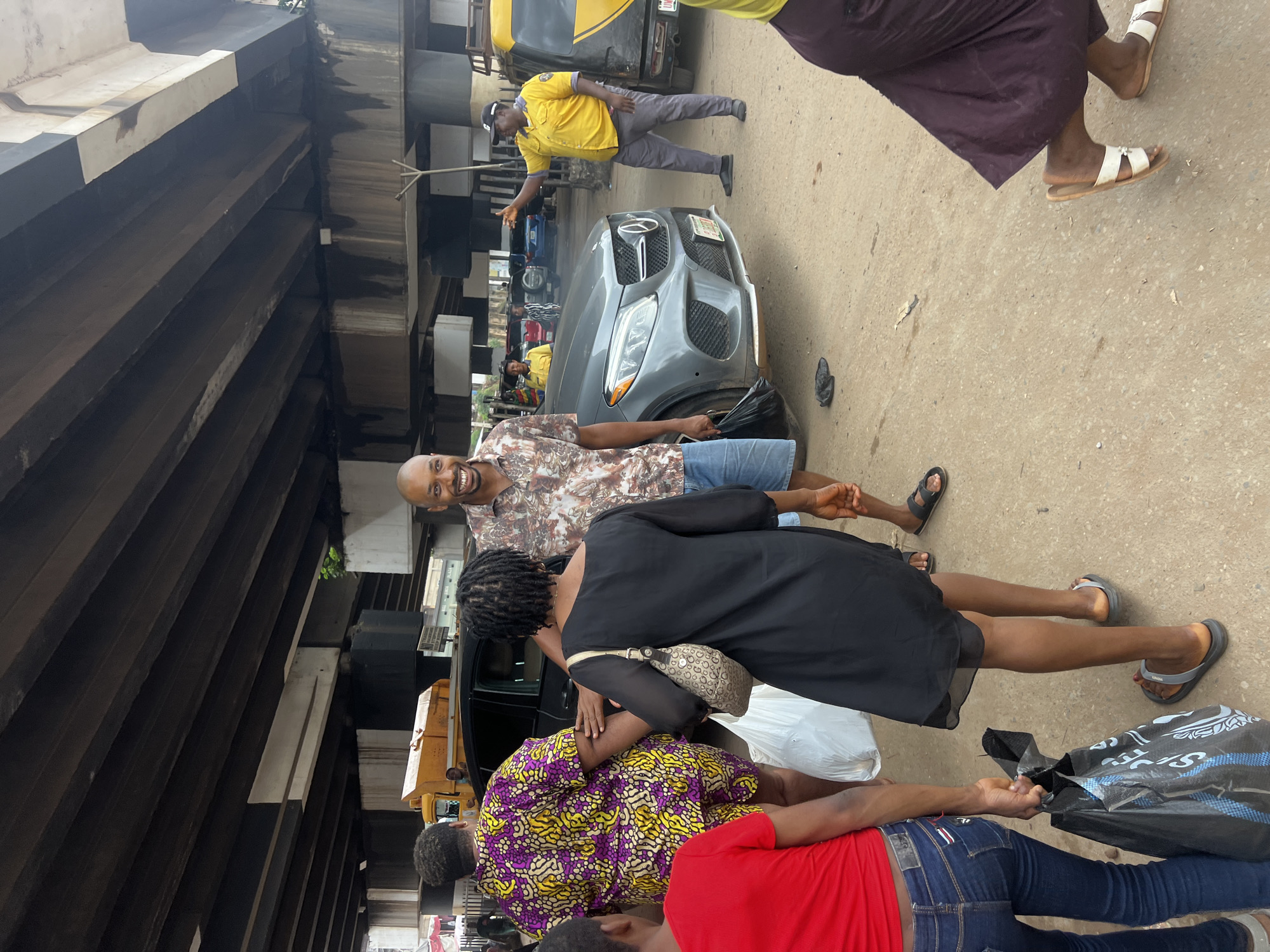 In Nkpor, Nigeria, under an overpass, a group of people are gathered on a dusty road next to a gray Mercedes-Benz. A man in a patterned short-sleeved shirt and denim shorts smiles at the camera, holding a black plastic bag. To his left, a woman in a black dress and a patterned yellow and purple outfit are talking. The woman in the black dress carries a large white plastic bag and a beige handbag. To the right, a man in a yellow uniform and a black cap, possibly a traffic warden, gestures with his hand, standing near a yellow and black vehicle. The environment suggests a busy, urban setting.