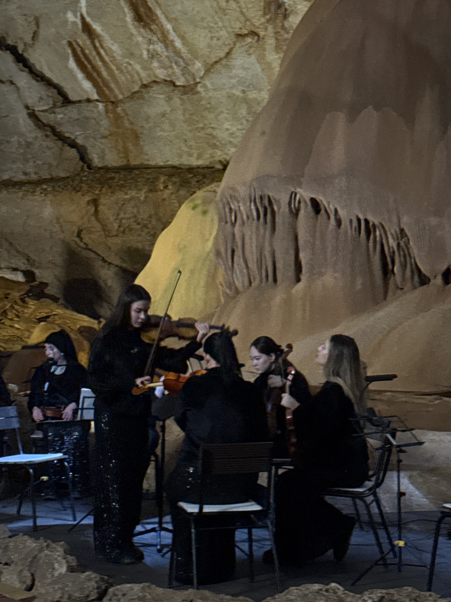 An ensemble of musicians plays violins inside a cave. The cave walls are textured with earthy tones and rock formations. Four women in black attire are visible; three are playing violins, and one is partially obscured on the far left, also holding a violin. They are seated on chairs, with music stands present. The lighting is dim, suggesting the performance is taking place in a natural, cavernous environment.
