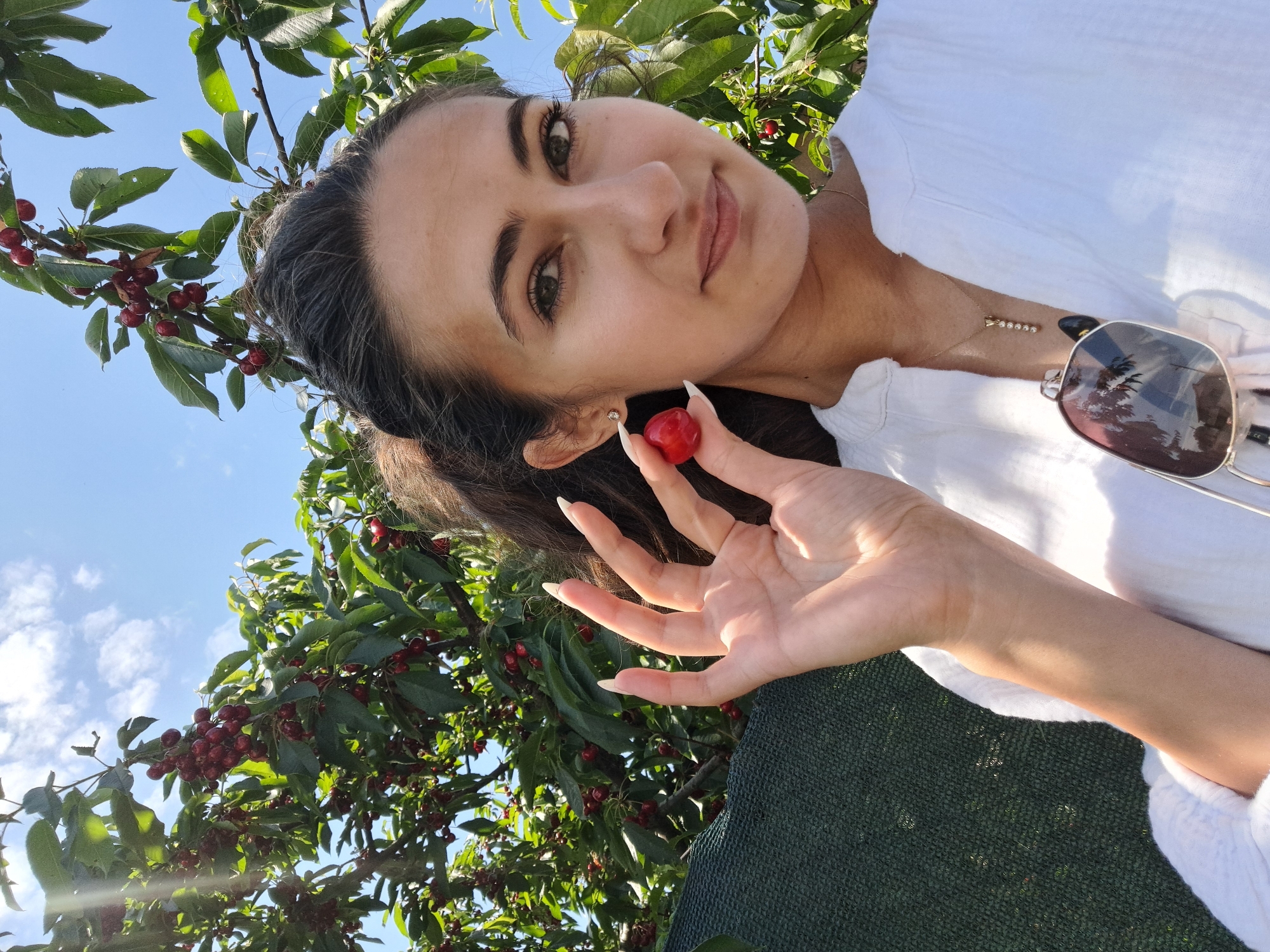 This is a selfie taken outdoors in Lokorsko, Bulgaria, likely during a sunny day, as indicated by the bright sunlight and clear blue sky with scattered clouds. The subject is a young woman with dark hair and brown eyes, wearing a white shirt. She is holding a single red cherry between her fingers, which appears to have just been picked from a nearby cherry tree. The tree, laden with ripe red cherries and green leaves, fills a significant portion of the frame, suggesting the photo was taken during cherry season.
The woman is looking towards the camera with a slight smile, her expression conveying a sense of joy and contentment. Her long, manicured fingernails are visible as she holds the cherry. A gold necklace with a delicate pendant and a pair of sunglasses are visible on her person. The background also shows a dark green mesh-like material, possibly a net or fence. The overall scene evokes a feeling of a pleasant summer day spent enjoying fresh fruit.