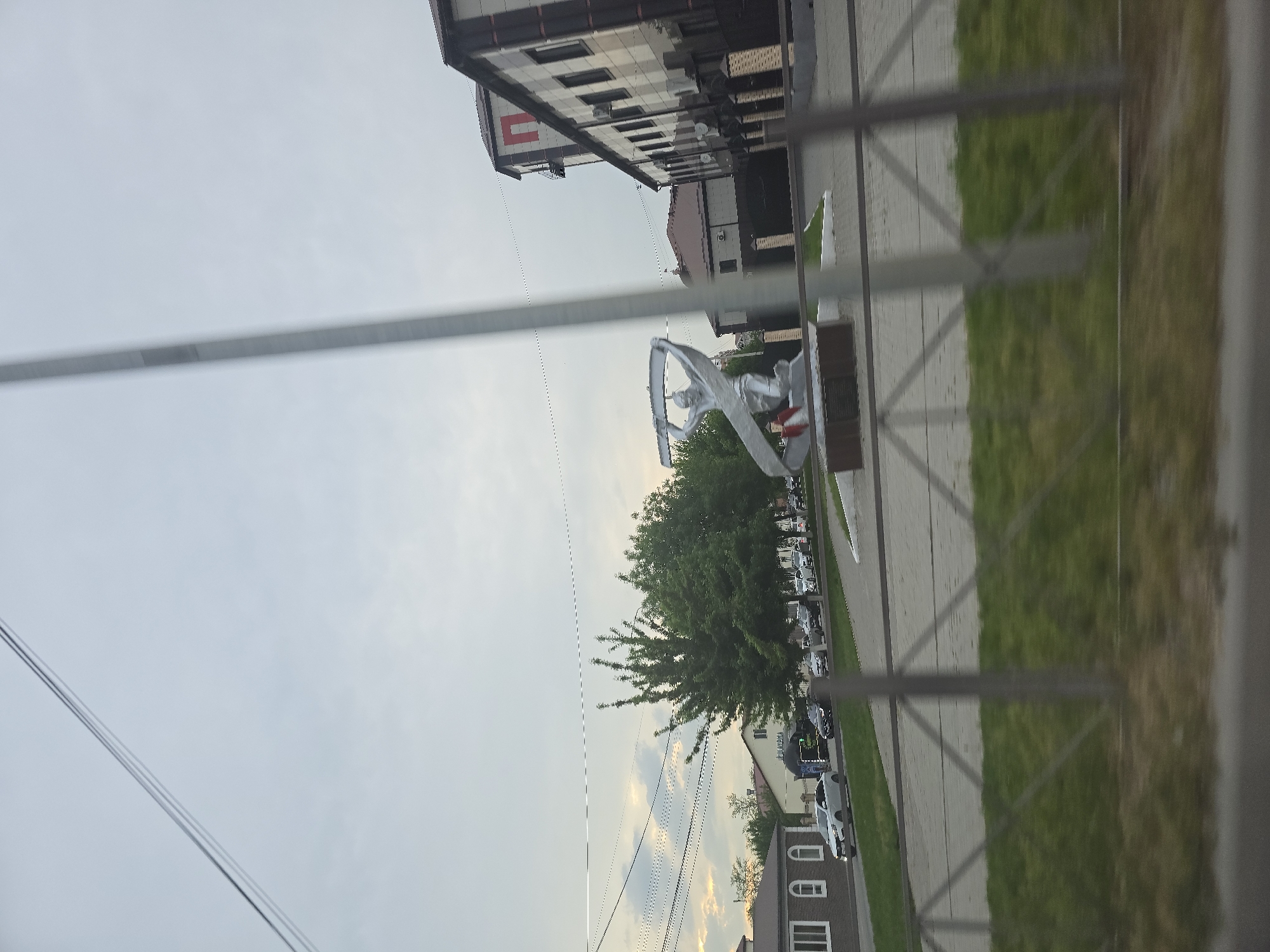 The image shows a street scene in an urban area of the city of Unknown, Unknown, taken from a low angle. A silver statue of an angel or winged figure is the focal point, positioned on a paved area next to a green grassy strip. Behind the statue, there is a tree and a street with parked cars. A building with multiple stories and windows is visible in the background, adjacent to the statue. Above, the sky is overcast with a mix of gray and light patches. A metal railing or structure is in the foreground, partially obstructing the view. The image is rotated such that the elements appear vertically arranged, although the actual scene is horizontally aligned.