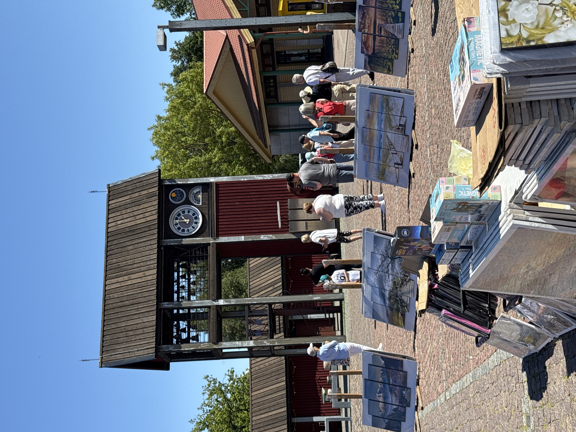 This is a photograph taken outdoors on a sunny day in Ventspils, Latvia. In the foreground, several canvases are displayed, showcasing landscape paintings. Some canvases are propped up on wooden stands, while others are stacked on a table with boxes of what appear to be art supplies. The canvases depict scenes with mountains, water, and wooden walkways.
In the middle ground, a group of people are gathered, appearing to be tourists or shoppers. Behind them stands a tall wooden structure resembling a bell tower, adorned with clocks and other circular decorative elements. To the right of the bell tower, a building with a yellow and red facade and a red roof is visible, possibly a shop or information center. The ground is paved with reddish-brown cobblestones. The background features green trees under a clear blue sky.