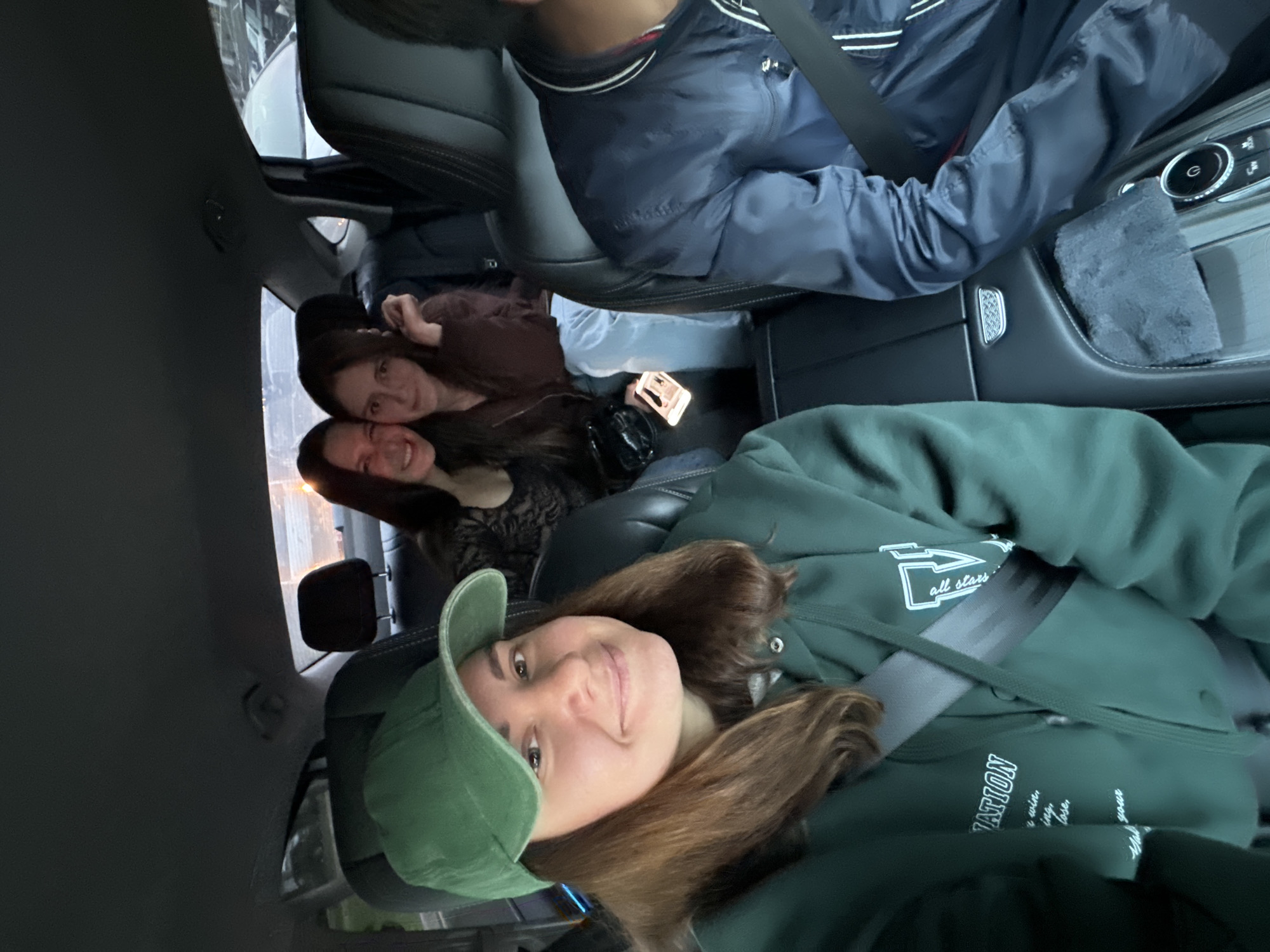 Four individuals are inside a vehicle. A person wearing a green hooded sweatshirt and green cap is in the foreground. Behind them are two women, and in the front passenger seat is someone wearing a navy blue jacket. All occupants seem to be wearing seatbelts. A mobile phone is visible in the background. The location is unknown.