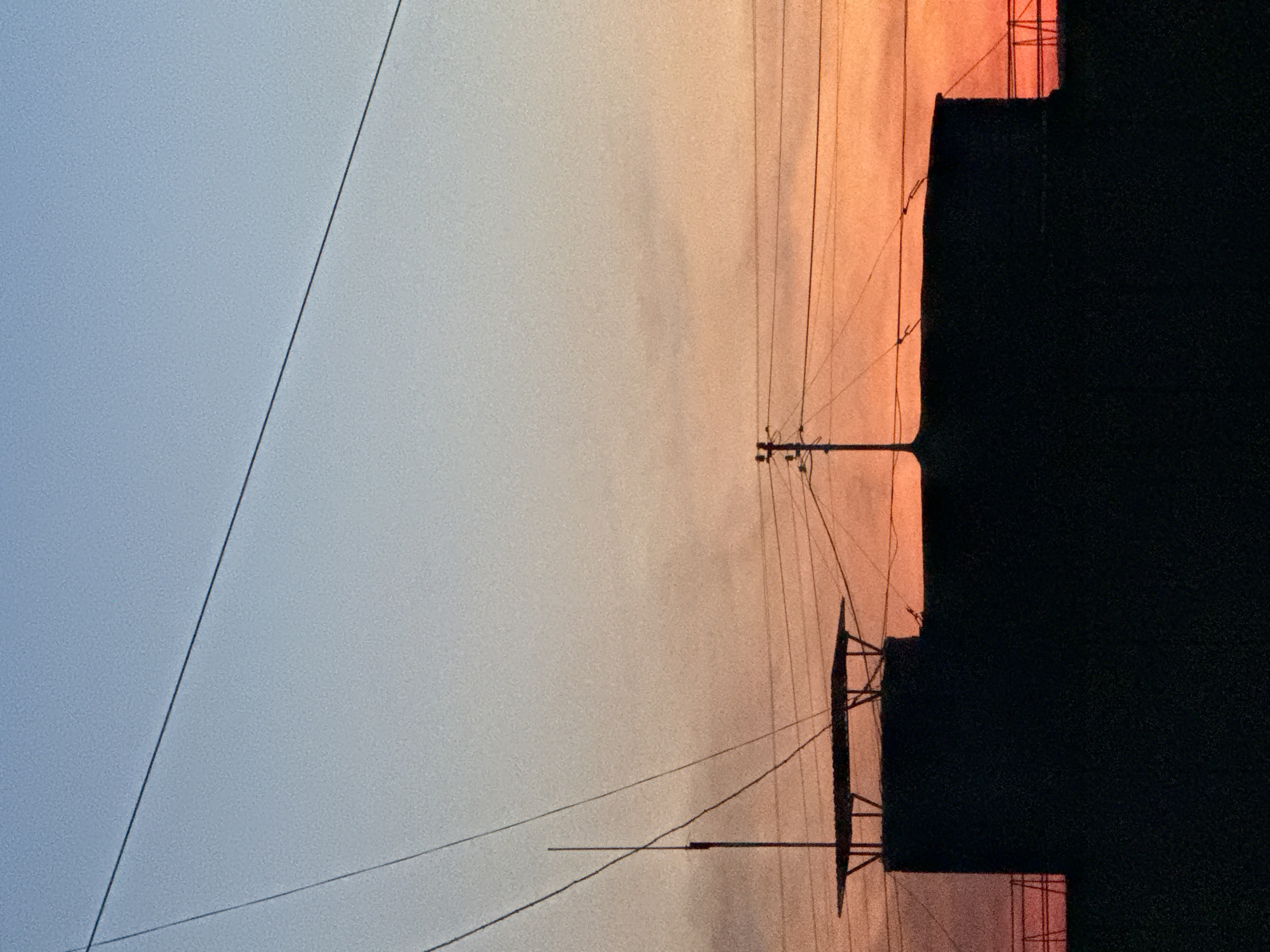 The image depicts a cityscape at dusk or dawn, possibly in Chaikovsky, Russia. The sky transitions from light blue to orange. Power lines extend across the sky, connected to utility poles. Silhouettes of buildings are visible against the lighter sky. There are no visible people or actions in the frame.