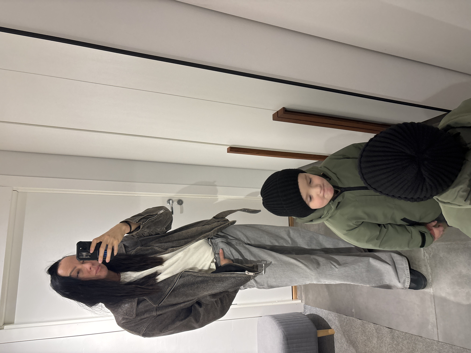 A woman takes a selfie with a child in a room that appears to be an entryway or dressing area. The woman is wearing a brown leather jacket, a white shirt, and light grey pants. She holds her phone up to the mirror, capturing the image. The child stands to her right, wearing a green jacket and a black knit hat. The room features white walls with dark trim, a wooden shelf, and a grey ottoman-like stool. The floor is grey. There's no definitive information suggesting that the image was taken in the city of Chaikovsky, Russia.