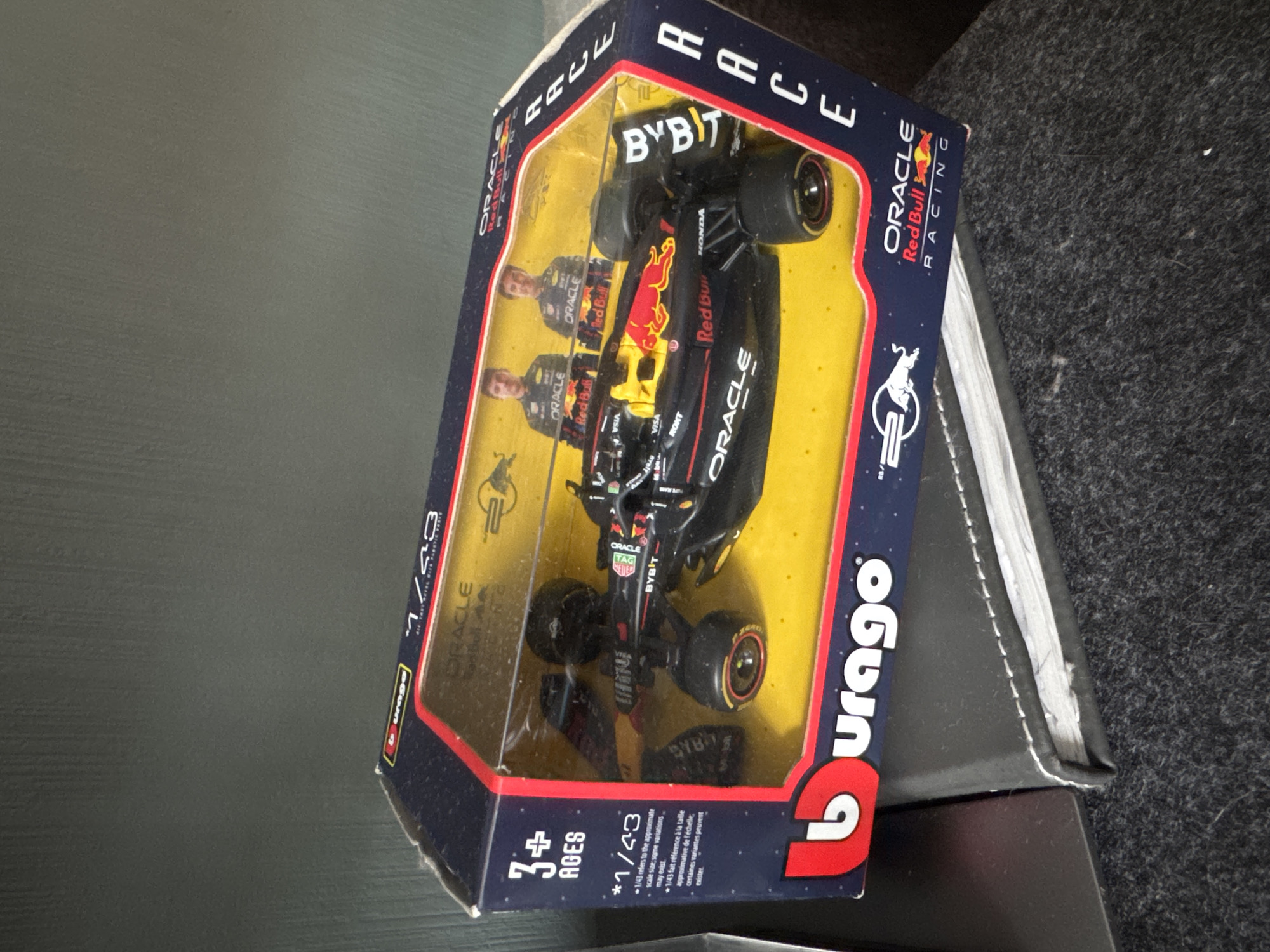 The image shows a Burago brand 1/43 scale model of the Oracle Red Bull Racing Formula 1 car in its packaging. The box is blue with red accents, and the car is black with red and yellow details. The packaging features the drivers and sponsors of the Red Bull team. The box is resting on a black object next to a surface with a dark, textured material.