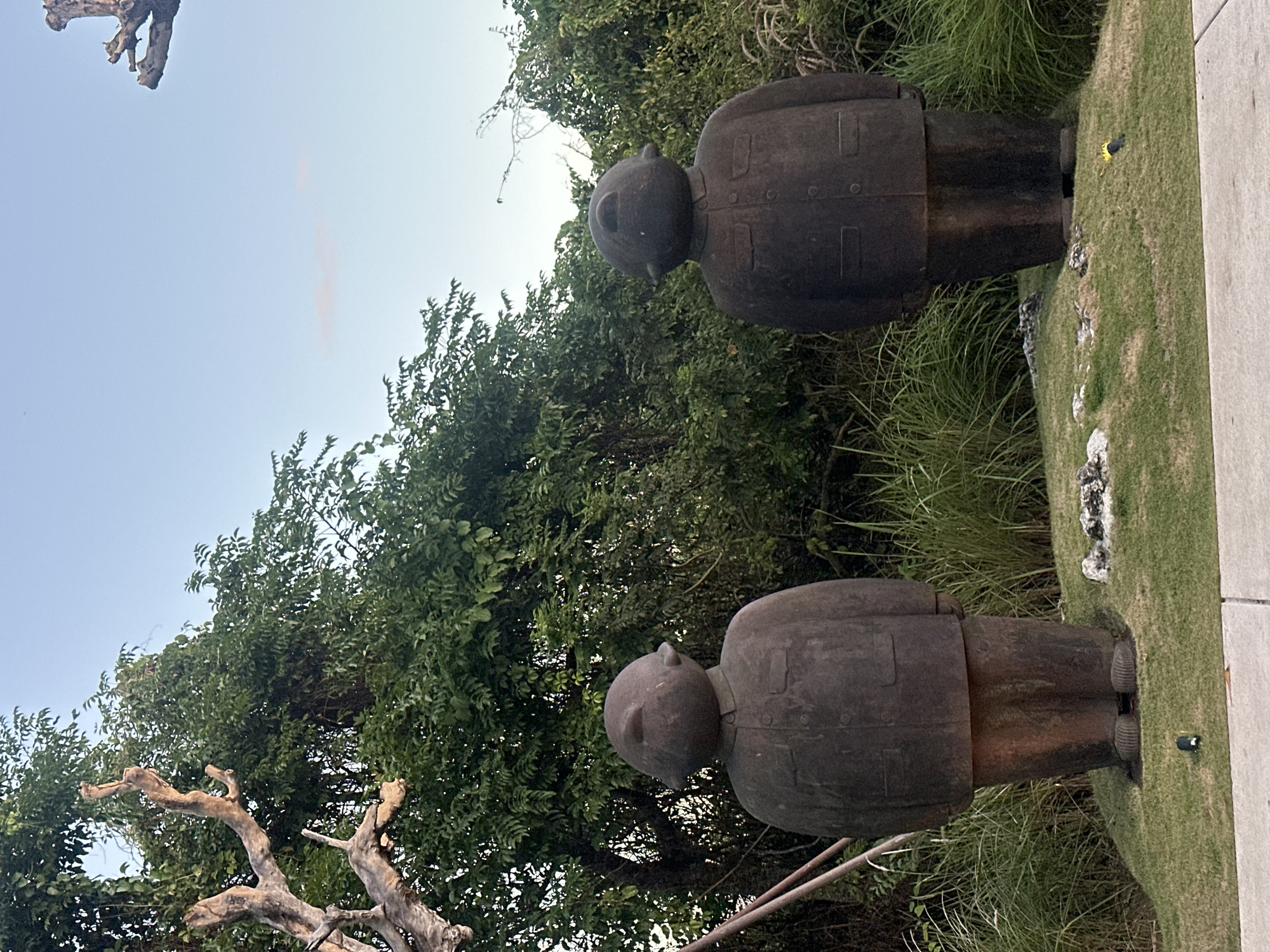 The image shows two dark, stone statues of men standing side-by-side on a grassy mound. The statues are roughly cylindrical in shape with rounded heads and appear to be wearing jackets with buttons and pockets. They are positioned in front of dense green foliage and a clear sky. In the foreground, there is a paved surface. The statues are located in Kuta Selatan, Indonesia.