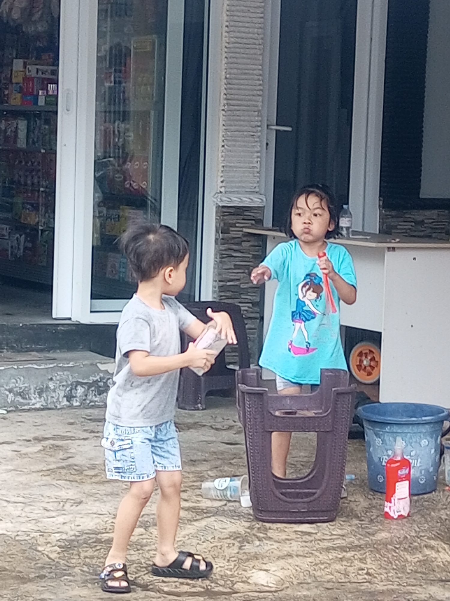 Two young children, a boy and a girl, are standing on a wet, textured outdoor ground surface in front of a building with a glass storefront. The boy, on the left, is wearing a grey t-shirt and light blue denim shorts. He is holding a clear plastic bottle with a pink cap in his right hand. The girl, on the right, is wearing a light blue t-shirt with a graphic of a dancer on it and grey shorts. She is holding a red and clear plastic toy in her right hand, and her left arm is extended forward. In the background, a dark brown woven plastic chair or bin is positioned between the children. To the right of the bin, a large blue bucket is visible, and next to it, a red bottle with white text. The building behind them has a white door frame and a dark-tinted glass entrance revealing shelves with products inside. The ground appears to be made of concrete with a rough, uneven texture. The lighting suggests it is daytime. The location is identified as Krasak, Indonesia.