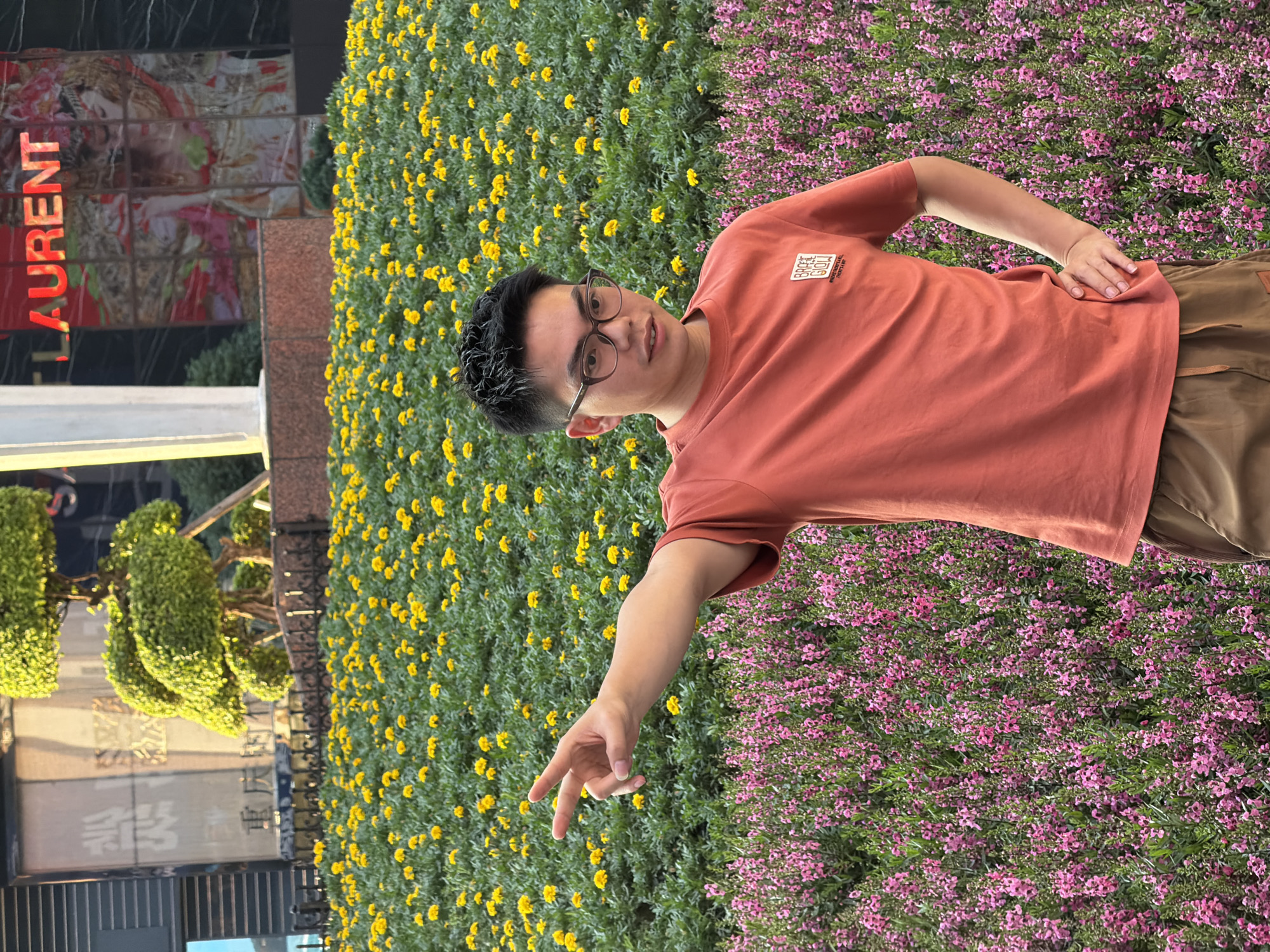 The image shows a young man standing in front of a vibrant display of yellow and pink flowers. He is wearing a coral-colored t-shirt with a square graphic and brown pants. The man has short, dark hair and is wearing glasses. He is looking towards the camera with a slight smile, and his left hand is extended, with his index and middle fingers pointing outwards.
The background is filled with dense greenery and blooming flowers, with yellow flowers in the upper left portion and smaller pink flowers in the lower right. Behind the flowers and to the left, a building with signage can be seen. The word "LAURENT" is prominently displayed in red lettering on the building. There are also decorative, topiary-like plants visible.
The lighting suggests it is daytime, possibly late afternoon given the warm tones. The overall scene conveys a sense of pleasant outdoor leisure. The man's expression is relaxed and friendly. The image appears to be taken outdoors in an urban environment, likely a public space decorated with flowers. The presence of the building with "LAURENT" text suggests a commercial or upscale area in Yuzhong District, China.