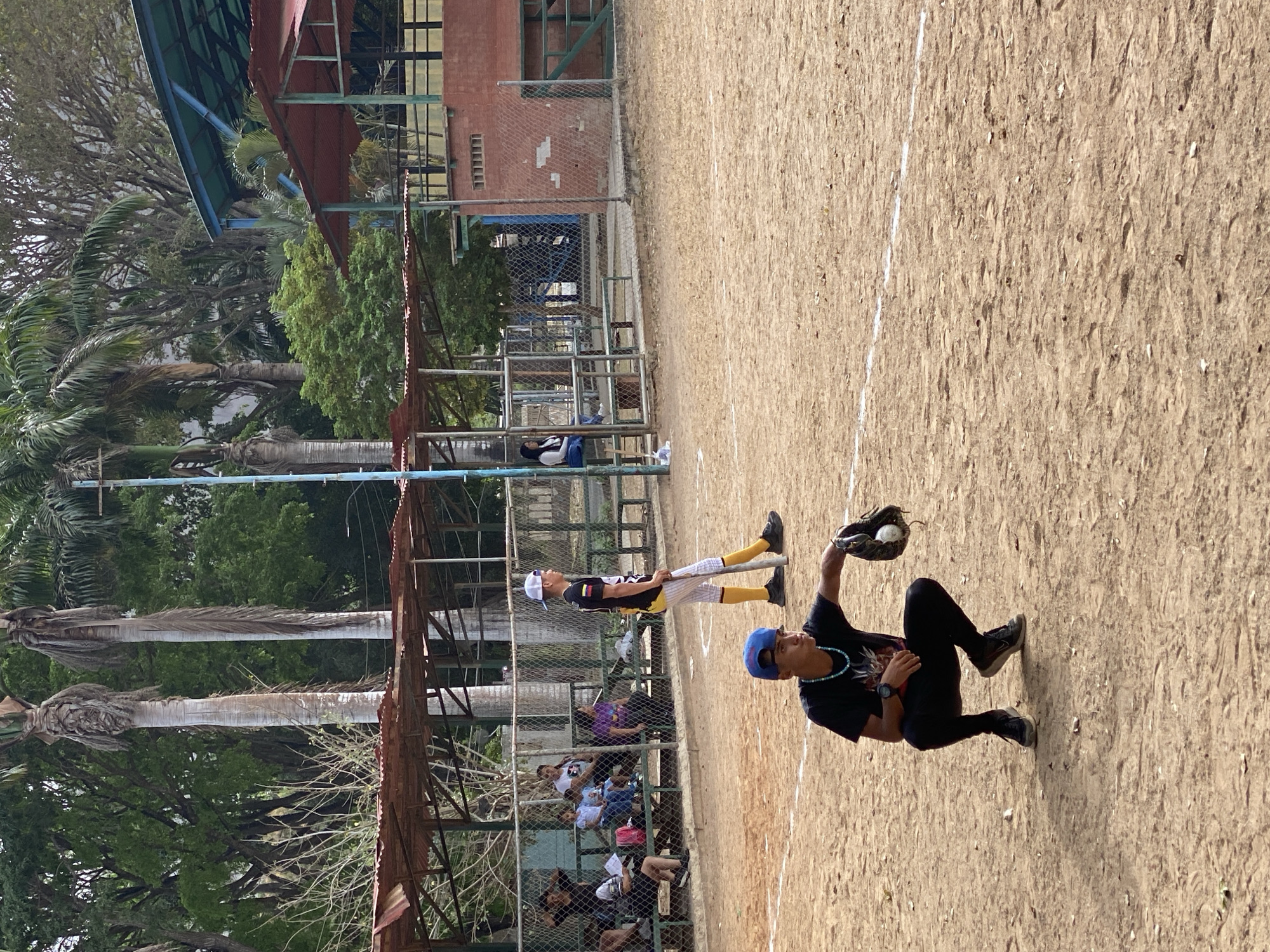 A youth baseball game is in progress. A young male in black pants and a black t-shirt with a blue baseball cap is crouching on the infield dirt, catching a baseball in his mitt. Another youth in baseball uniform stands in the background. In the background, a chain-link fence separates the field from a spectator area with a rusted metal awning covering a bench. Several spectators are visible. Outside the fence are trees. The location is Las Piedras, Venezuela.