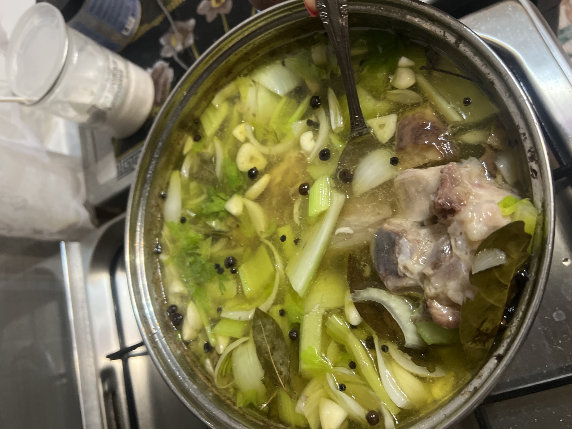 A close-up, overhead shot captures a large pot filled with a simmering broth. The pot contains an assortment of ingredients, including chunks of meat, celery stalks, sliced onions, garlic cloves, bay leaves, and whole peppercorns. The broth itself appears light yellow and is actively bubbling, indicating it is hot. Two spoons are partially submerged in the liquid, one with its handle extending out of the pot, suggesting the broth is being stirred or ingredients are being sampled.
In the background, out of focus, a glass jar with a metal lid is visible. The jar contains a white granular substance, possibly salt or sugar, and has a label with some text on it, though it's largely illegible. The overall setting suggests a kitchen environment, with hints of cookware and what appears to be a stovetop. The lighting suggests it could be daytime, but there are no strong indicators of the time of day. The scene is domestic and practical, focusing on the preparation of food. The location context of Krasnoe-na-Volge, Russia, adds a specific geographical setting to this everyday culinary activity. No people are clearly visible in the frame, only the presence of hands holding the spoons.
