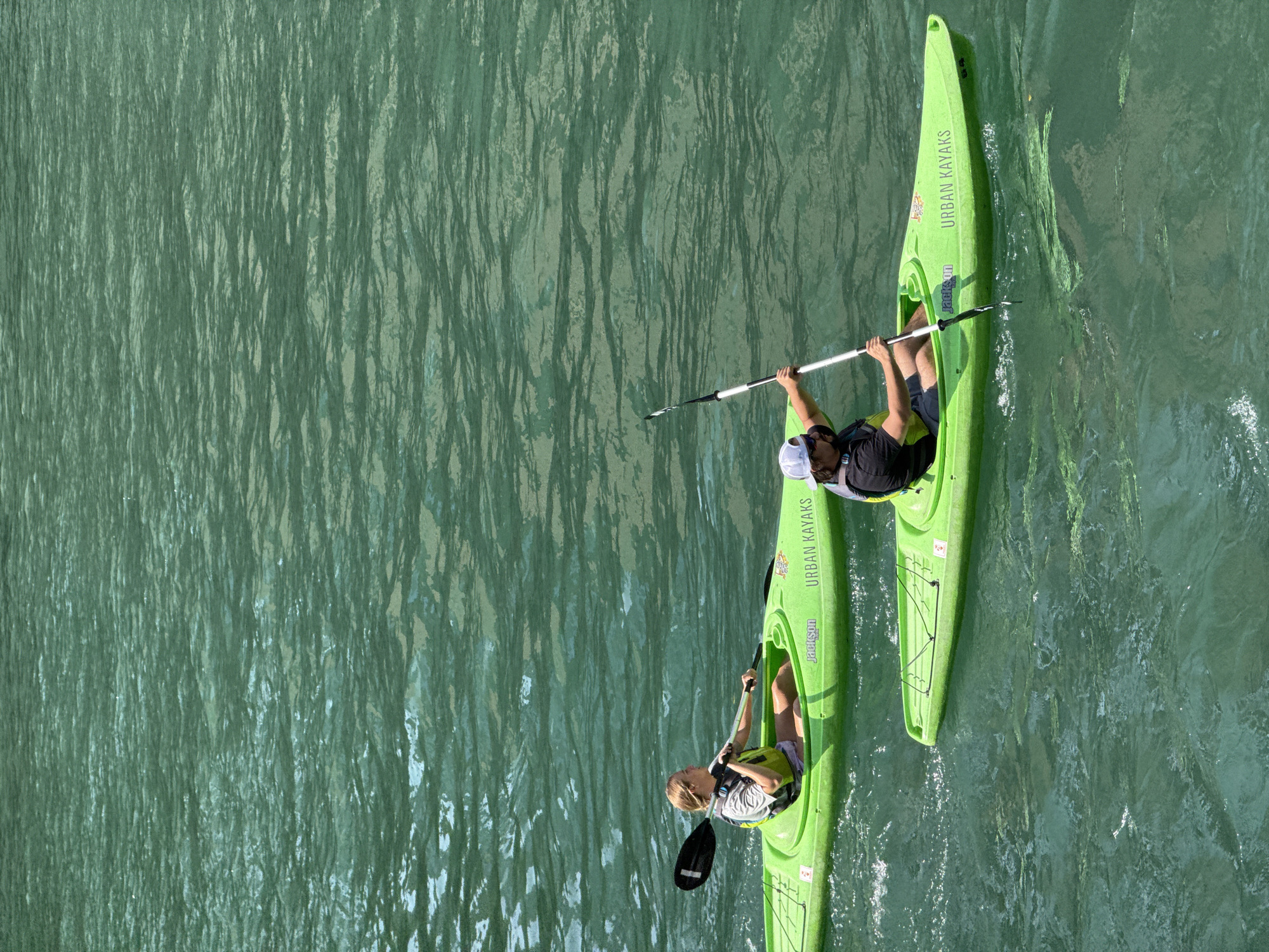 Two individuals are kayaking on a body of water that appears to be a river or lake. The water is a vibrant turquoise color with ripples and reflections of sunlight. Both kayaks are bright green and have the text "URBAN KAYAKS" printed on the side.
In the foreground, a person wearing a black t-shirt, dark shorts, a white baseball cap, sunglasses, and a yellow life vest is paddling a kayak. They are holding a double-bladed paddle, using it to propel themselves forward.
Behind them and to the left, another person is kayaking in a similar green kayak. This individual is wearing a grey t-shirt and a yellow life vest. They are also holding a paddle.
The overall scene suggests a recreational activity on a clear, sunny day. The location is not identifiable as being in Chicago from the provided image.