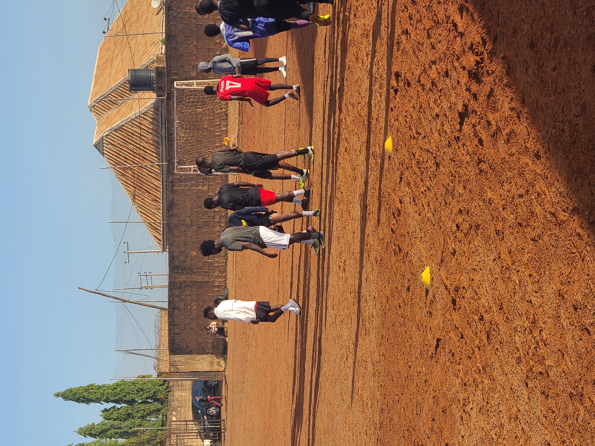 A group of young men is seen on a reddish-brown dirt sports field under a clear blue sky. They appear to be engaged in or preparing for a sports activity, likely soccer or football, suggested by their athletic attire, the presence of two yellow training cones on the ground, and a goal net visible in the background.
There are approximately 10-12 individuals, mostly dark-skinned, standing in various positions across the field. Many are wearing athletic clothing, including jerseys, shorts, and sports shoes. One person in a red jersey has "P.JAL" and the number "17" visible on their back. Another has "Es" partially visible on a black jersey. Their long, distinct shadows stretch across the dusty ground, indicating bright sunlight and suggesting it is midday or early afternoon.
In the background, several buildings with corrugated metal roofs and brick walls are visible, alongside a chain-link fence, utility poles, and tall, conical trees. A dark car is also parked near the edge of the field. A black water tank can be seen on the roof of one of the buildings. The overall scene depicts an outdoor sports environment in a sunny, possibly warm, setting, characteristic of a semi-urban or rural area.