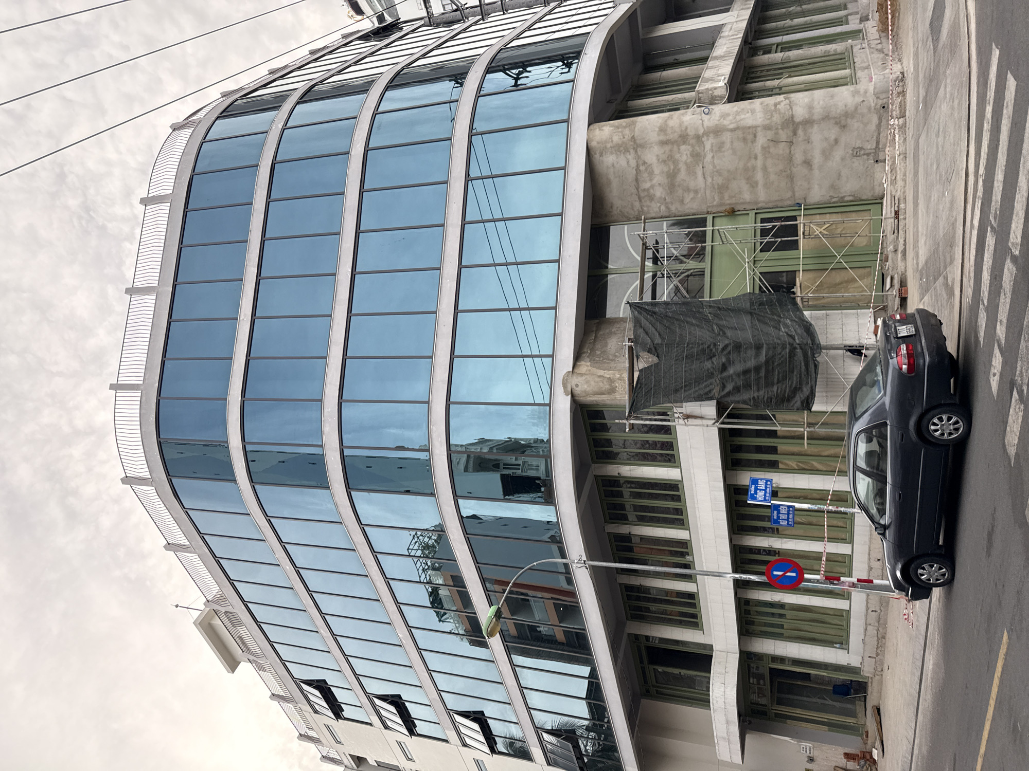 The image shows a modern, curved building with large blue-tinted windows on a cloudy day in Nha Trang, Vietnam. The building appears to be under construction, with concrete sections and scaffolding visible on the lower floors. A dark gray sedan is parked on the street in front of the building, near a pedestrian crossing. A lamppost stands on the sidewalk, and a traffic sign with a red circle and a diagonal bar is also present. In the background, power lines are visible against the sky. The street is paved with asphalt.