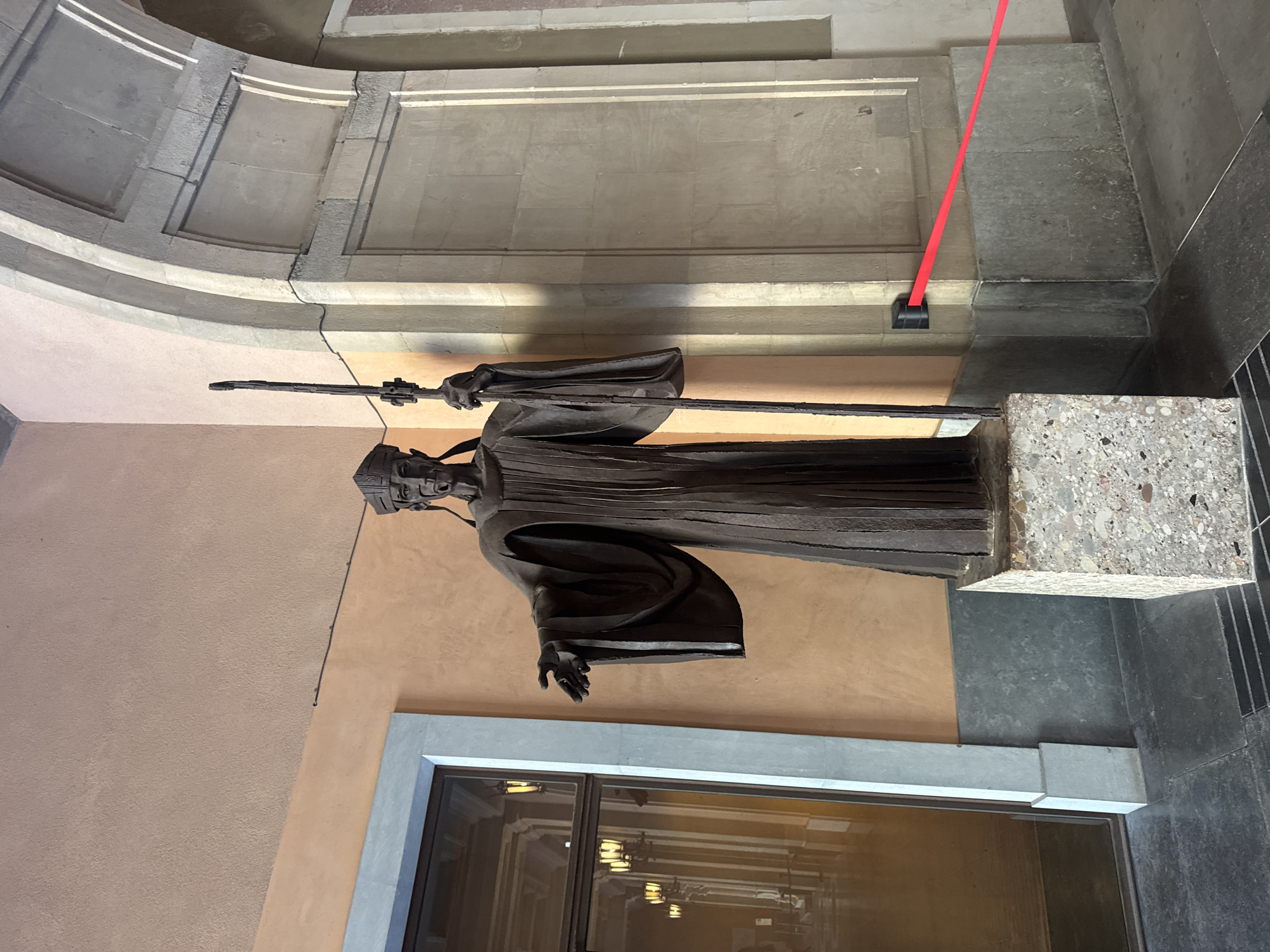 A bronze statue of a robed figure wearing a headdress stands on a speckled stone pedestal. The statue is positioned outdoors in what appears to be a covered walkway or portico. The figure holds a long staff in its right hand and has its left hand extended forward. The background features a light peach-colored wall and a stone archway. To the left, a large glass door reveals a reflection of an interior hallway with lampposts. A red barrier tape is visible in the lower right portion of the image, attached to the wall. The ground is paved with dark tiles. This statue is located in the city of Monistrol de Montserrat, Spain.