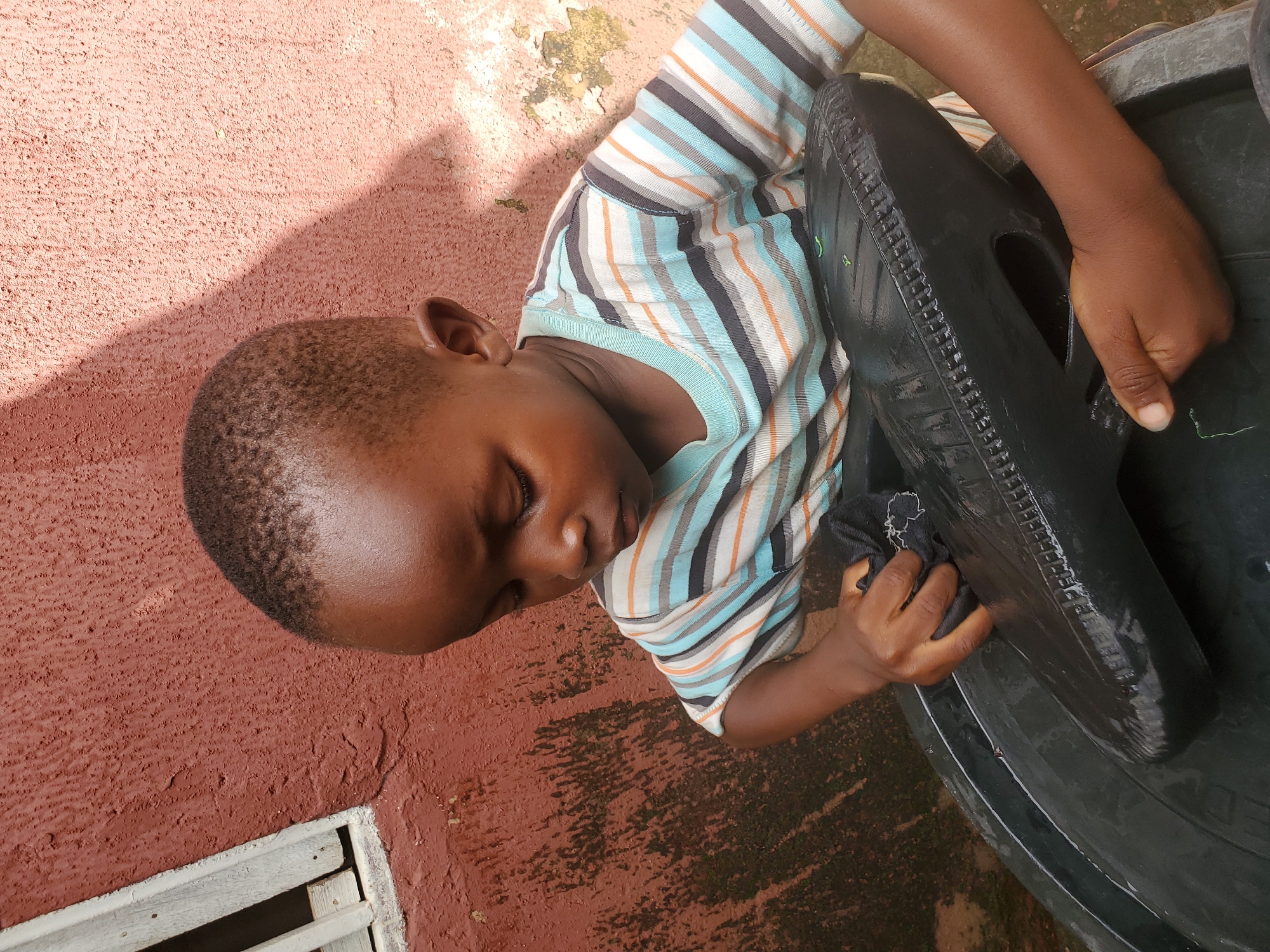 A young Black boy, appearing to be around three years old, is shown outdoors, possibly in the city of Jos, Nigeria. He is looking down and interacting with a large, black, possibly plastic object, which could be a tire or a container. He is wearing a striped t-shirt with blue, orange, and gray horizontal stripes. The background consists of a textured, reddish-brown wall, which appears to be weathered and shows some signs of dampness and moss growth on the left side. There is also a portion of a white-painted wooden structure, possibly a window frame, on the upper left side of the image. The lighting suggests it is daytime, with a shadow cast on the wall behind the boy. The boy's hands are engaged with the black object, one hand holding a dark cloth. The overall scene is candid and captures a moment of play or interaction with the object and the cloth.
