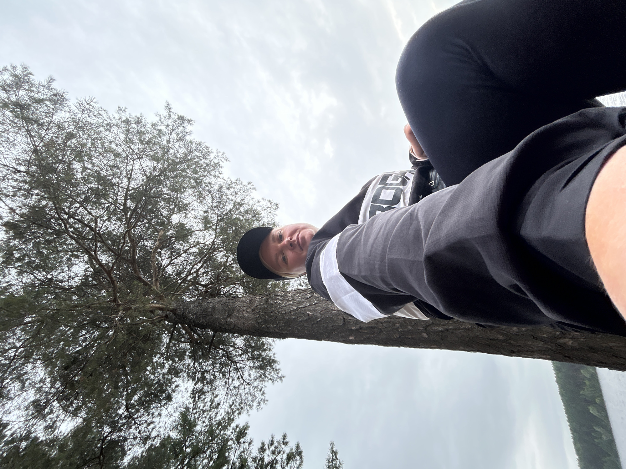 The image is a low-angle, wide-shot selfie taken outdoors. The primary subject is a person, appearing to be Caucasian with light-colored hair, wearing a black baseball cap, a black and white long-sleeved shirt, and black pants. The person is looking directly at the camera, with a neutral expression.
The setting appears to be a natural environment, with a large tree trunk prominently featured in the foreground, extending diagonally across the frame. The tree has rough bark and sparse foliage. Behind the tree and the person, the sky is overcast with clouds, suggesting a cloudy or possibly rainy day. In the distance, on the right side of the image, a body of water and a treeline are visible, with dense evergreen trees.
The activity appears to be a casual moment, possibly a pause during an outdoor activity like hiking or exploring. The low angle of the shot gives a sense of looking up from a resting position. The lighting is diffused due to the cloudy sky.
Visible text on the person's shirt includes "ROGO" in large, bold white letters, with what appears to be a smaller "G" or similar character above it. The overall mood is calm and natural. The location context provided is Chaikovsky, Russia, which aligns with the natural, possibly northern European landscape. The time of day is not explicitly indicated but the overcast sky suggests it could be daytime, perhaps late afternoon or morning.