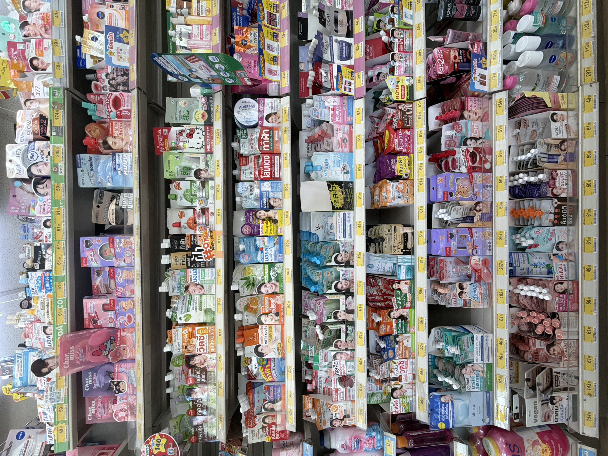 The image shows a retail display shelf filled with various beauty and skincare products. The products are arranged in an organized manner on multiple shelves, with some items placed in boxes and others individually displayed. The shelves are well-lit, highlighting the colorful packaging of the products. There are no people visible in the image. The overall environment appears to be a retail store, possibly a pharmacy or a department store, given the assortment of health and beauty items. The presence of Thai language on the product labels suggests the location is in Thailand, specifically in Ko Samui District. The products include items like facial masks, serums, creams, and other cosmetic goods.