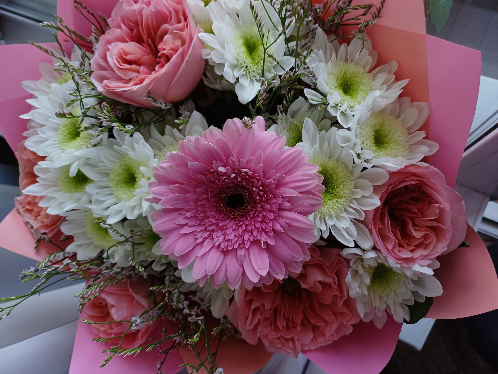 A close-up, top-down shot showcases a vibrant bouquet of flowers. The arrangement features a prominent pink Gerbera daisy in the center, surrounded by white chrysanthemums with green centers. Several soft pink roses are interspersed throughout, some with tightly furled petals and others more open. Delicate sprigs of small purple and green filler flowers add texture and depth. The bouquet is wrapped in layers of bright pink and peach paper. The background is blurred, but hints of a light-colored surface and a dark wooden surface are visible. The lighting is soft and natural, highlighting the colors and textures of the flowers. The scene appears to be indoors, possibly near a window, given the diffused light.