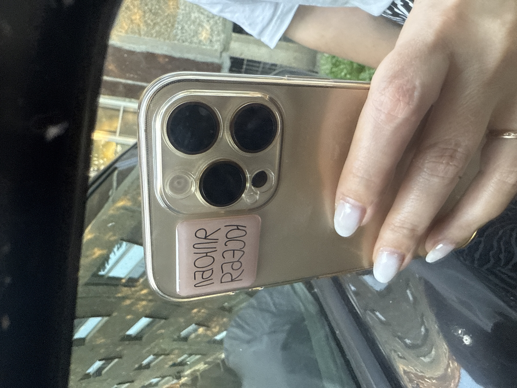 A close-up shot shows a person holding a gold iPhone with a clear case. A square pink sticker with black lettering is affixed to the back of the phone case. The lettering appears to be in a stylized font, with two lines of text. The upper line reads "NAOINR" and the lower line reads "RAAJION", possibly in reverse or a playful script. The phone is positioned in front of a window or glass, and the reflection of a building with multiple windows is visible. The background is slightly blurred, but suggests an urban outdoor setting. The image does not contain any information that would indicate its location in Chelyabinsk, Russia.