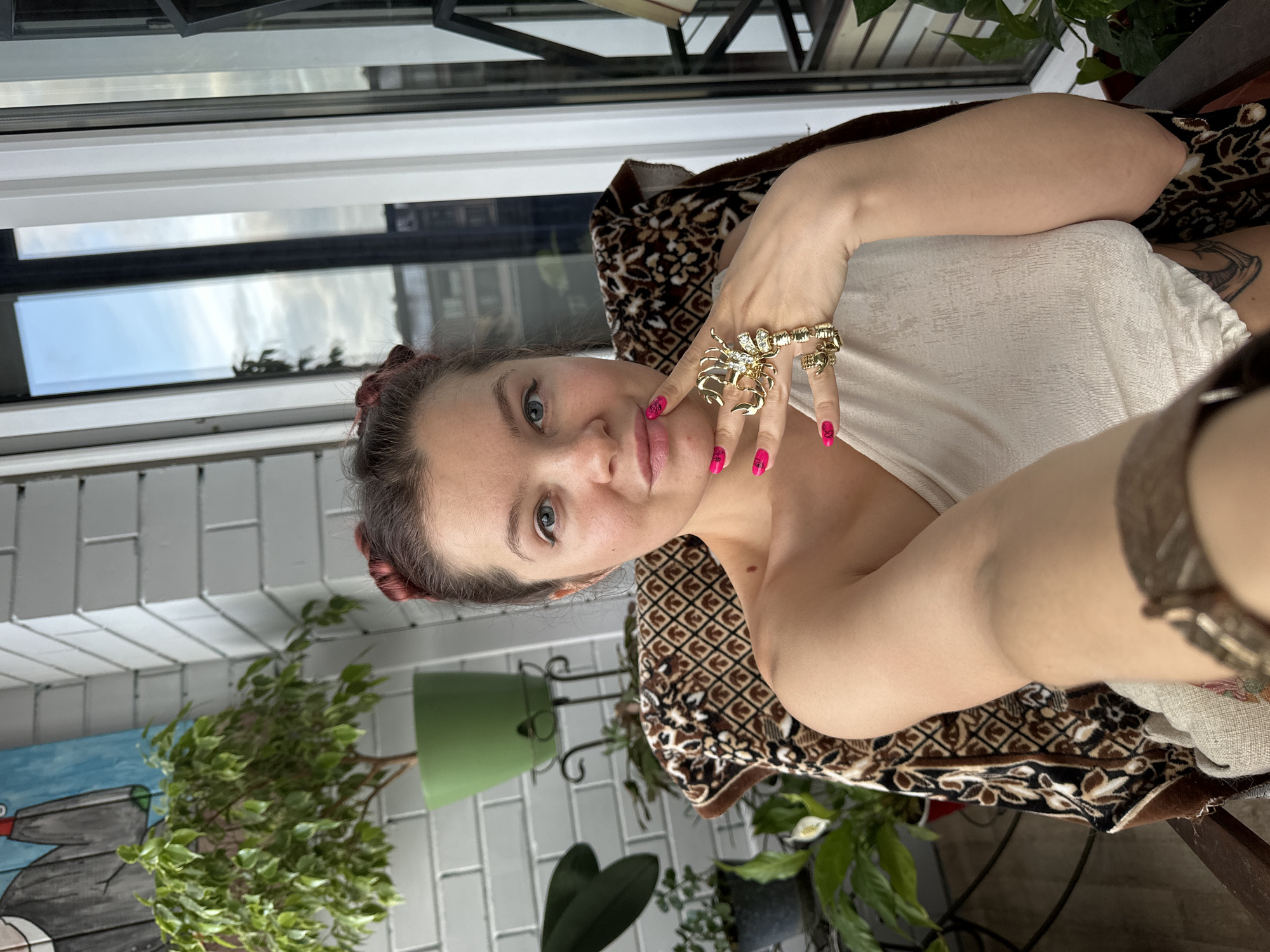 The image is a vertical, selfie-style portrait of a young adult, rotated 90 degrees counter-clockwise. The subject is a woman with light skin, blue-green eyes, and reddish-brown hair styled in a bun or pulled back, partially visible on the top and left of her head. She wears a light-colored, possibly off-white, textured top. Her fingernails are painted bright pink with small black designs.
A prominent feature is the elaborate, gold-toned ring shaped like a scorpion, extending over several fingers of her right hand. Another gold ring is partially visible on her left hand, and a matching gold-toned bracelet is seen on her right wrist. She is leaning against a patterned fabric, possibly a blanket or chair upholstery, featuring brown, black, and cream geometric and organic motifs. A tattoo is partially visible on her right upper arm.
The background suggests an indoor setting, likely a sunroom or enclosed balcony, illuminated by natural daylight. Behind her is a large window or glass door, reflecting a partially cloudy sky and what appears to be distant greenery or buildings. Several potted plants with green foliage are scattered throughout the background, one in a distinct green pot. A white tiled wall is visible behind the plants on the left. On the far left, a piece of artwork depicting a stylized grey and black animal (possibly a bird or duck) on a blue background is partially visible. The person gazes directly at the camera with a neutral expression. No visible text is present in the image. The time of day appears to be daytime, with soft, natural light. This scene is located in Vladimir, Russia.