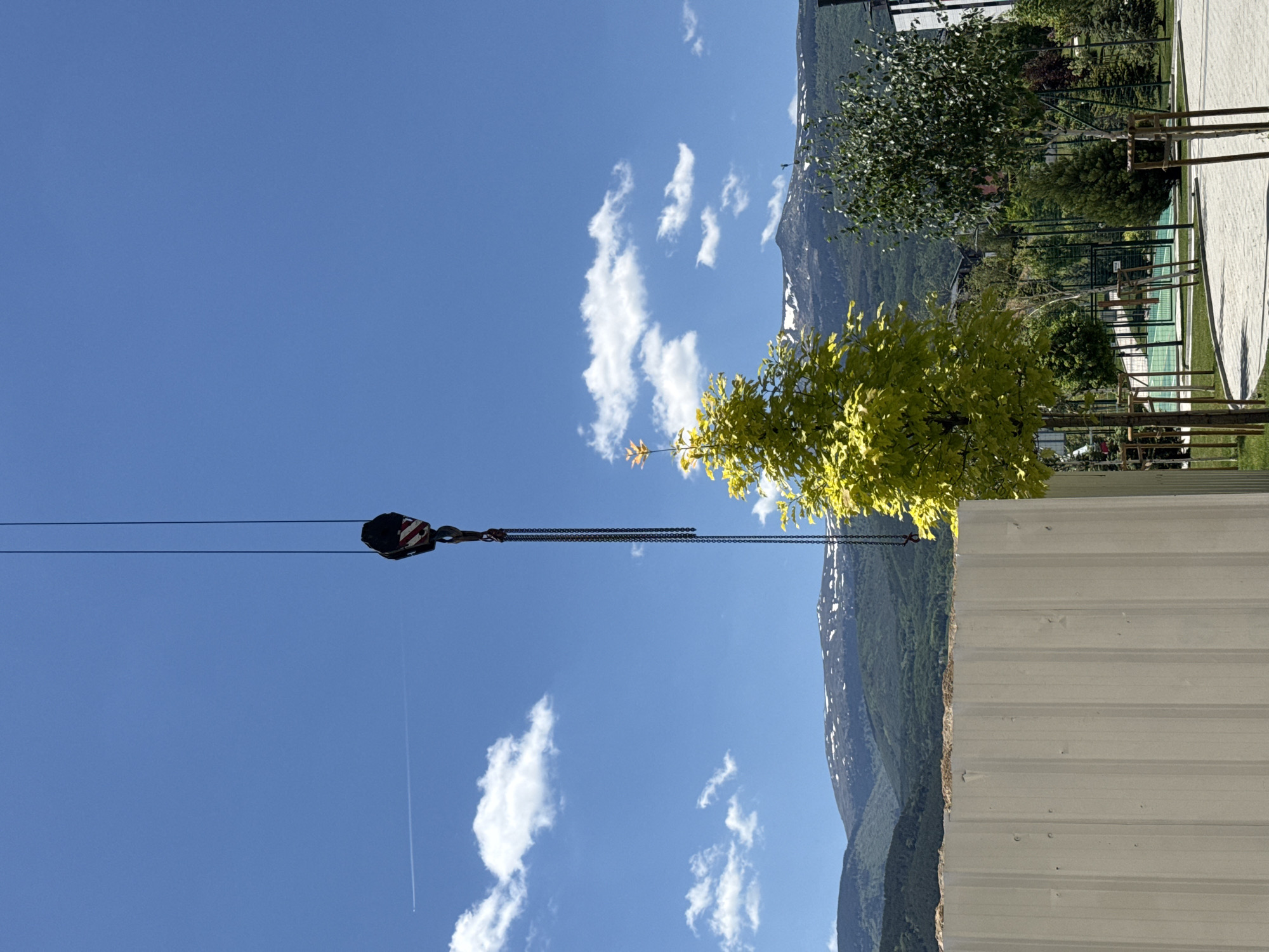 The image shows a blue sky with scattered white clouds. A crane hook with cables is visible in the upper left part of the image. A mountain range is present in the background. In the foreground, there is a tree with green and yellow leaves, as well as a building wall.
The location of the image is unknown and not Sofia, Bulgaria.