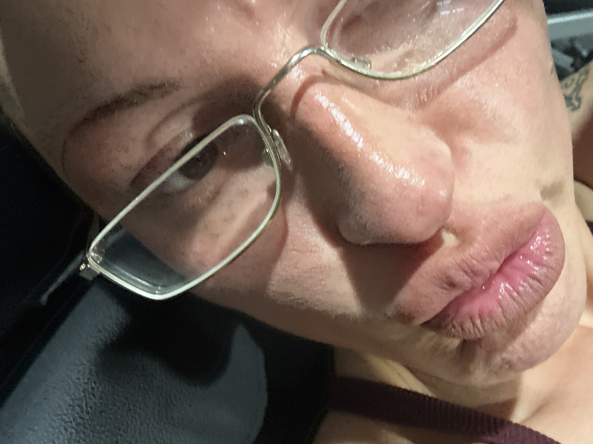 A close-up photograph focuses on a person's face, angled to the left. The subject is wearing glasses with silver frames, which partially obstruct the left eye. The person's skin appears to be flushed. A tattoo is visible on the upper right side of the image. A black object is in the lower-left corner of the photo, possibly a chair or seat. A maroon or dark red strap is seen near the bottom center of the frame, possibly clothing. The subject's lips appear slightly pursed or puckered. No information suggests that this image was taken in Tunbridge Wells, United Kingdom.