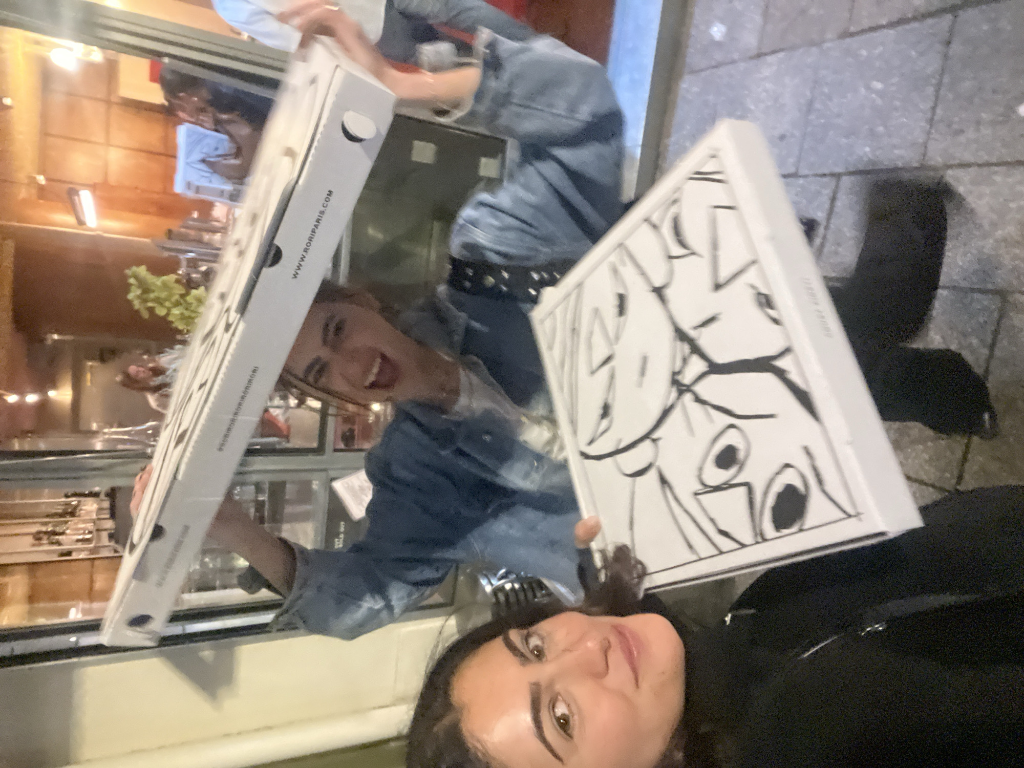 A selfie shows two women holding two white boxes. One box is emblazoned with a black and white drawing. The other box has the website "WWW.RORIPARIS.COM." The women appear to be on the sidewalk in front of a Parisian business.