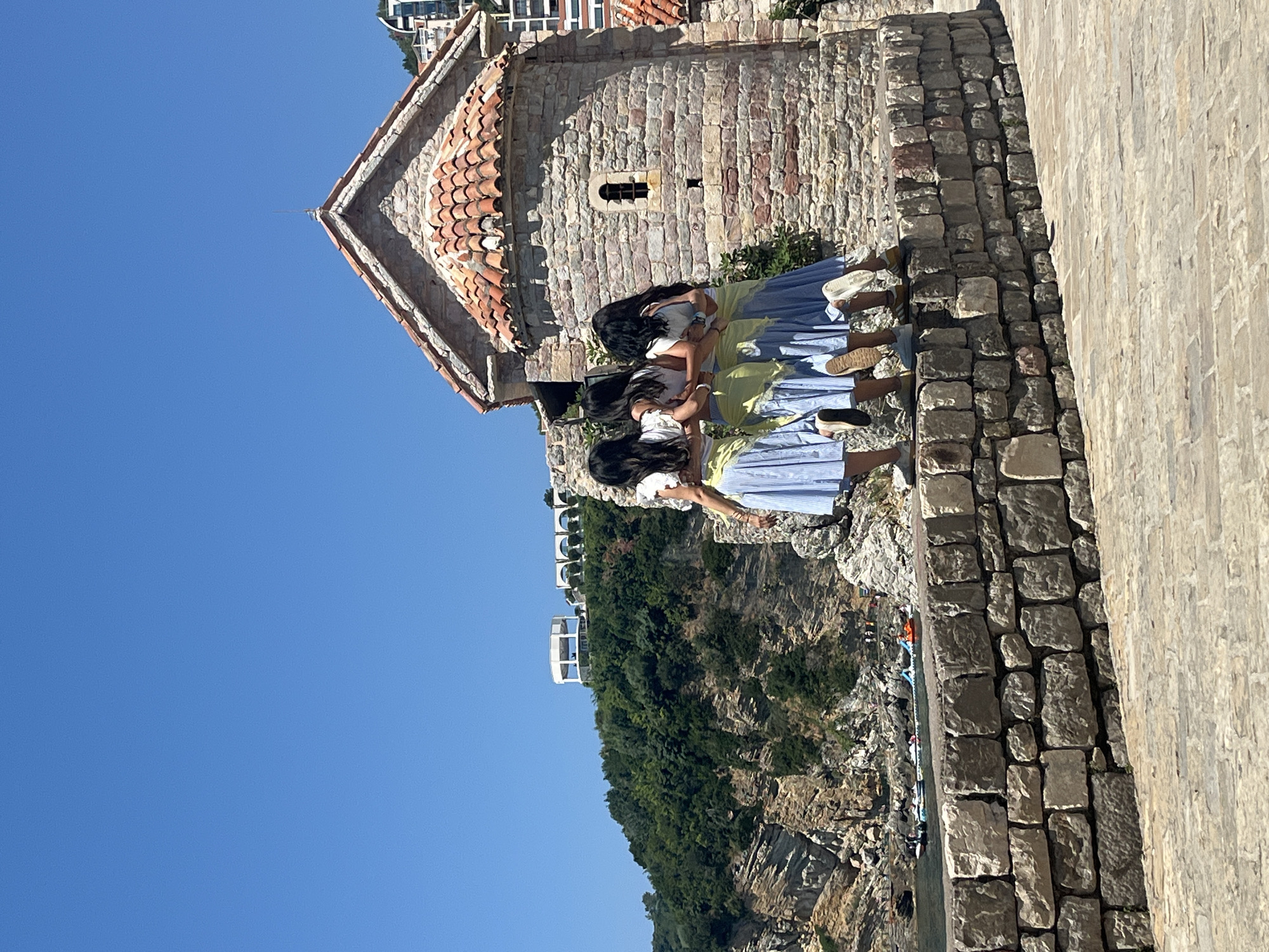 The image shows three young women from behind, huddled together. They are wearing light-colored dresses and appear to be looking down at something. The woman in the foreground is wearing a yellow and blue striped dress, the middle woman is in a lime green and blue striped dress, and the woman in the back is in a white and blue striped dress. They are standing on a stone wall, which is part of an ancient building. The building has a red tile roof and walls made of rough-hewn stone. Behind the building, a steep, verdant cliffside drops down towards the sea, where some boats are visible. The sky is a clear, bright blue, suggesting it is a sunny day, likely mid-morning or afternoon. The scene appears to be a historic site or a scenic viewpoint, possibly in Budva, Montenegro, given the characteristic stone architecture and coastal cliff landscape. The women's posture suggests a shared moment of curiosity or observation.