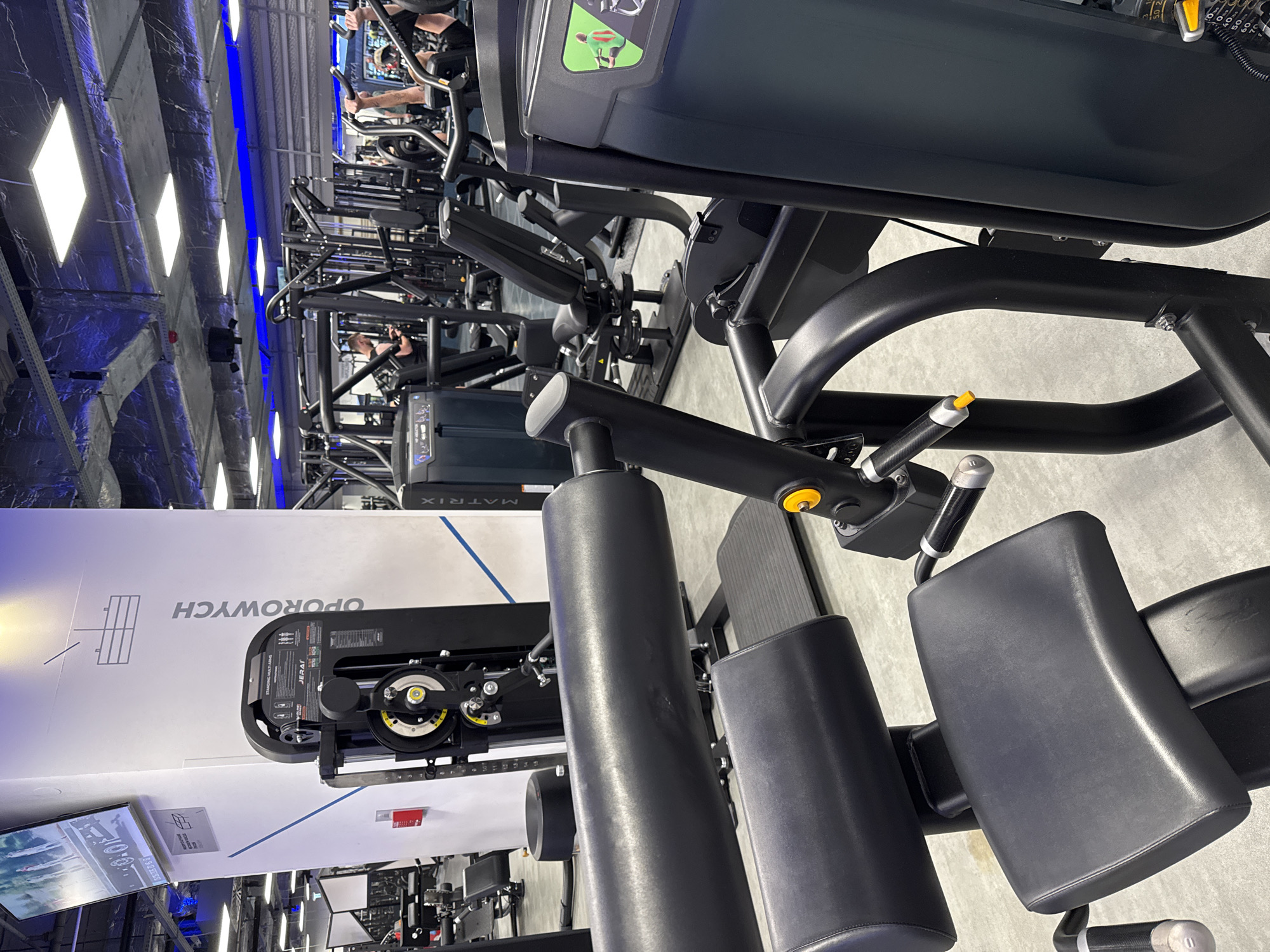 The image depicts the interior of a contemporary fitness center located in Unknown, Unknown. In the immediate foreground, the black upholstered seat, thigh, and shin pads of a gym machine are visible, featuring black matte metal framing and yellow adjustment knobs on its right side.
Centered in the midground, a white rectangular pillar displays a flat-screen television showing people in motion, possibly a workout video or commercial. Below the screen, the word "OPOROWYCH" is written vertically in gray capital letters, along with a schematic diagram of a cable pulley system. Adjacent to the pillar on the right, a black "JERAI STANDING MULTI ARMS" machine with yellow weight selection pins and instructional diagrams is identifiable. To the left of the pillar, a male individual in a black tank top and shorts is visible, appearing to be resting or engaging with a mobile device near a gym machine. Further into the midground on the right, another person is engaged in an exercise, with their legs elevated on a machine.
The background reveals an extensive array of black strength training and cardio equipment. The ceiling features exposed silver ductwork, square white LED panel lights, and linear blue LED strip lighting along the perimeter, creating a distinct ambiance. A black cylindrical speaker hangs from the ceiling in the upper mid-left. The floor is light gray. A "MATRIX" branded machine is partially visible on the far right. The overall scene is well-lit, indicating active use of the gym facilities.