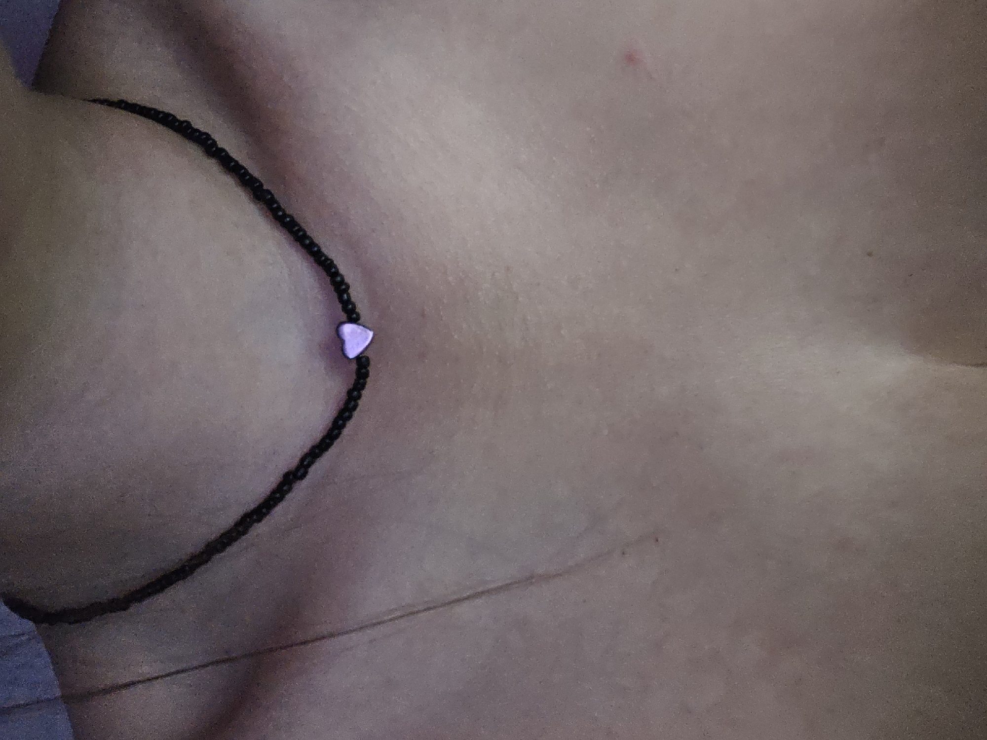 A close-up, waist-up shot shows the chest and neck of a person wearing a black beaded necklace with a small purple heart charm. The background is out of focus and appears to be a dark, possibly textured surface. The lighting is dim, creating a moody atmosphere. The image does not provide any information about the location in Petropavlivska Borshchahivka, Ukraine.