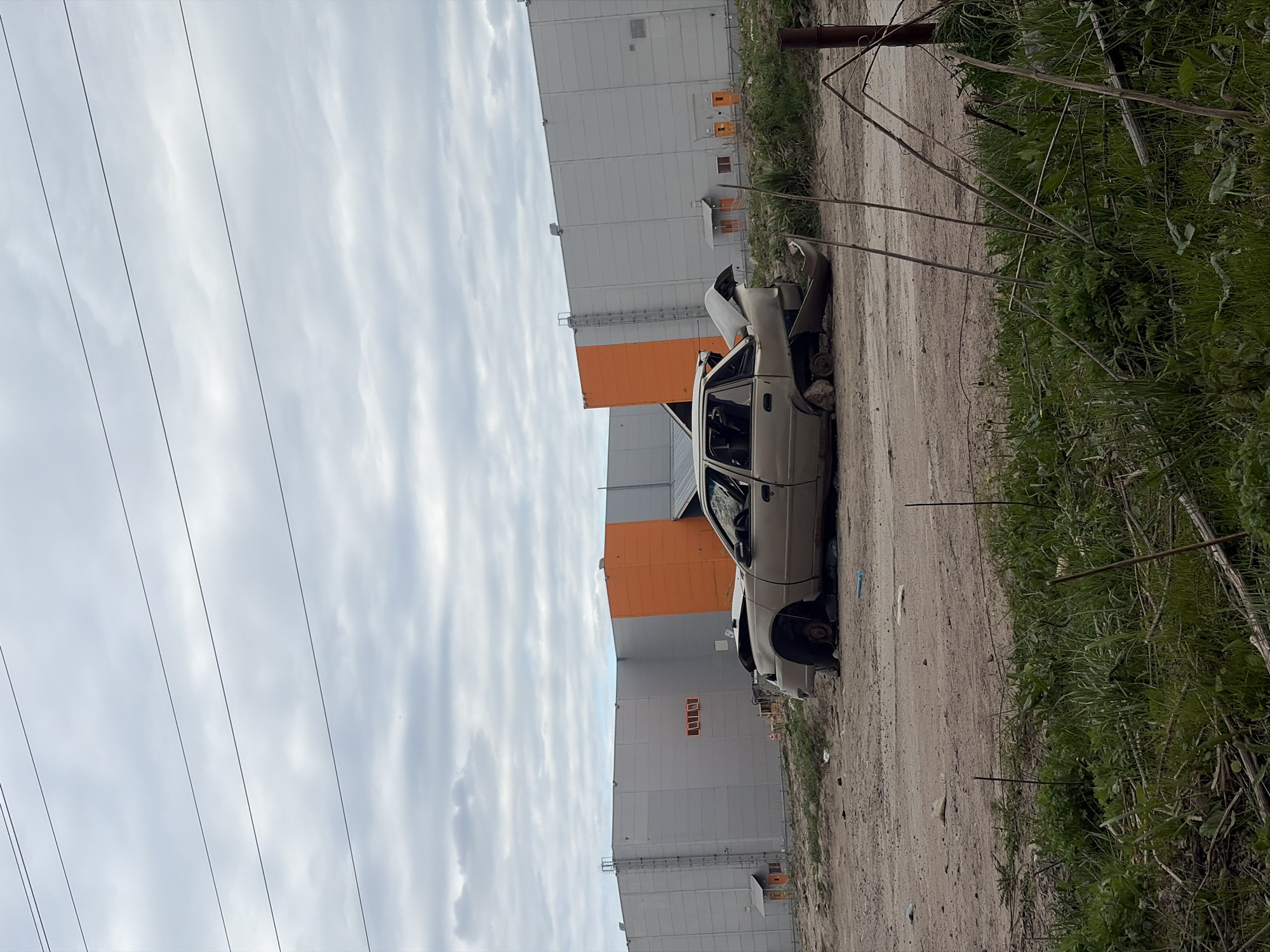 The image shows a wrecked, light-colored sedan on a dirt road. The car appears heavily damaged, with missing tires and a partially open hood. The road is bordered by overgrown vegetation on one side and a large, modern industrial building on the other. The building is primarily gray with orange accents and what appears to be the name "LERON" on the facade. Overhead are power lines against a cloudy sky. The setting suggests a neglected area, potentially an industrial outskirts in Saint Petersburg, Russia.