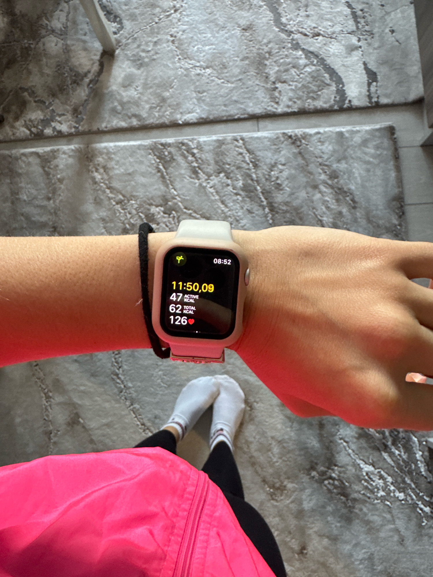The image shows a person's left arm and wrist, wearing a light-colored smartwatch with a black band. The smartwatch displays the time as 08:52, with activity metrics showing 11:50:09 duration, 47 active kcal, 62 total kcal, and a heart rate of 126 bpm. The person is wearing a bright pink jacket over black leggings and white socks. The background consists of a grey, marbled floor. The location is not specified in the image or its metadata.