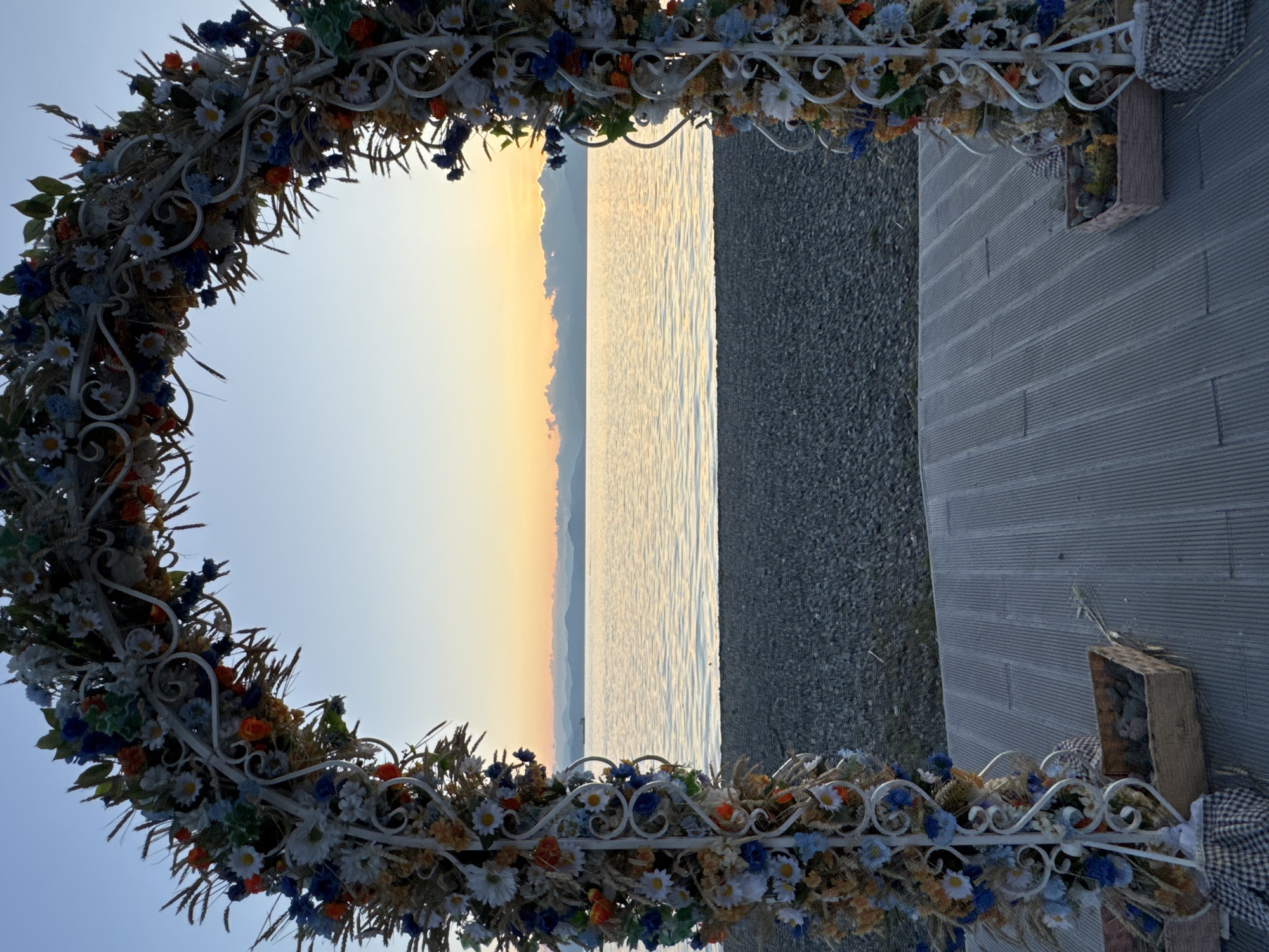 The image shows a wedding arch decorated with artificial flowers and wheat stalks set up on a wooden platform facing a body of water, likely the Avacha Bay in Petropavlovsk-Kamchatsky, Russia. The sun is setting, casting a warm glow over the calm water and silhouetting the distant mountains and a ship. The sky is a gradient of pale blue and soft orange, with a few scattered clouds. The arch is adorned with white, blue, and orange flowers, as well as dried wheat ears. In front of the arch, on the platform, are two woven baskets filled with what appear to be decorative stones or pebbles, and fabric drapes are visible at the base of the arch. The foreground shows a pebbled beach leading to the water.