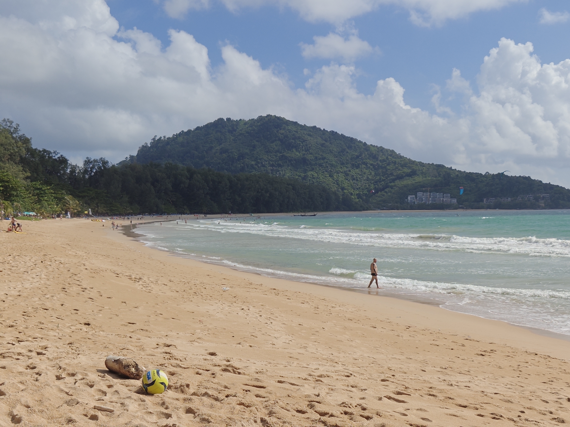 In the city of Sakhu, Thailand, a wide, sandy beach stretches under a partly cloudy blue sky. The foreground features a yellow and black volleyball and a piece of driftwood on the tan sand. Further down the beach, a man in black swimming trunks walks along the shoreline, his legs wet from the turquoise ocean waves. In the background, numerous people are scattered along the beach, some near palm trees and vegetation. Boats are visible on the water, and a lush, green, tree-covered hill forms the backdrop, with some buildings nestled into its slope. Kite surfers are also present in the ocean.